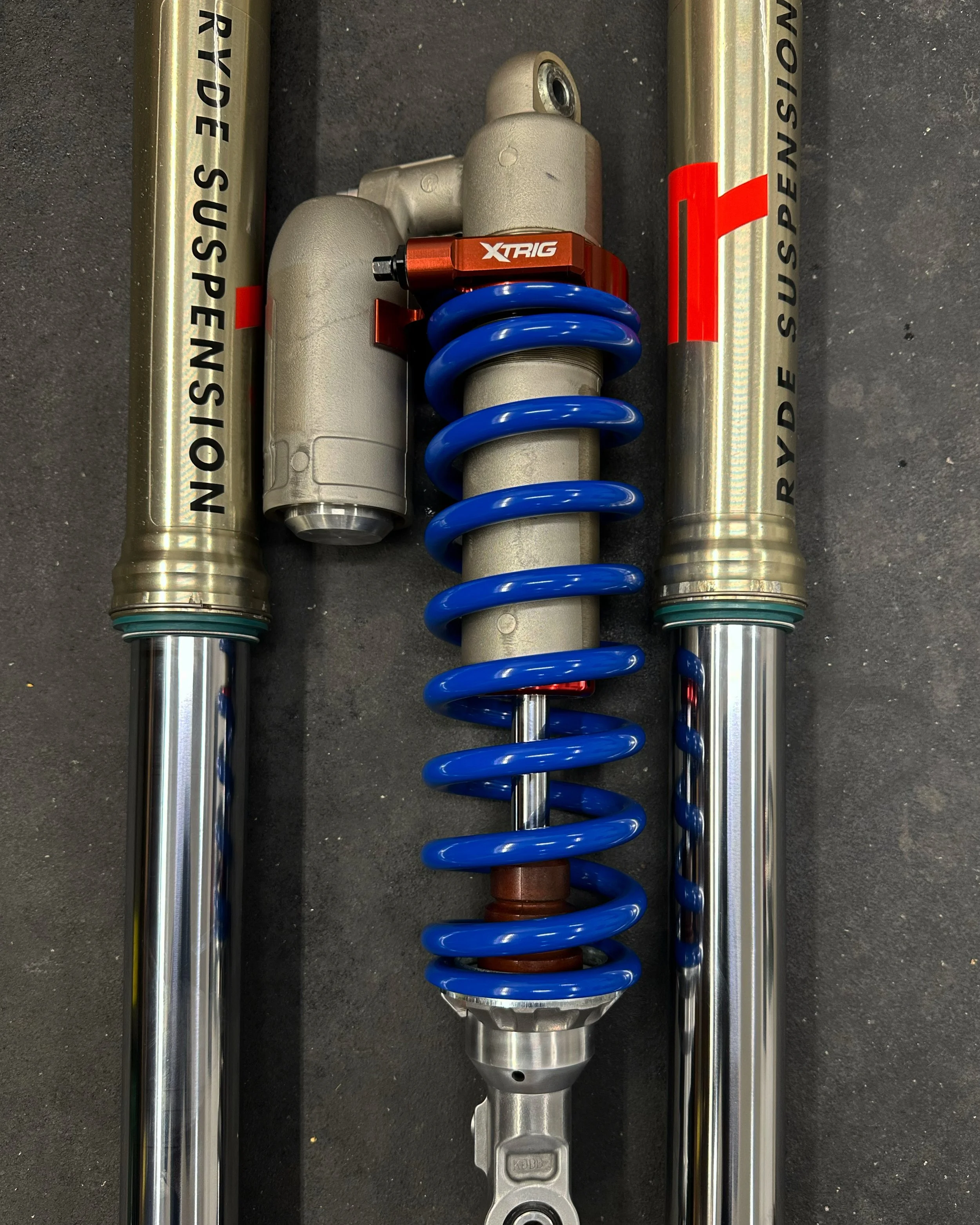 A close-up of motorcycle suspension parts including two gold-colored front forks with black text, a blue coil spring, and a silver shock absorber with red accents, placed on a black surface.