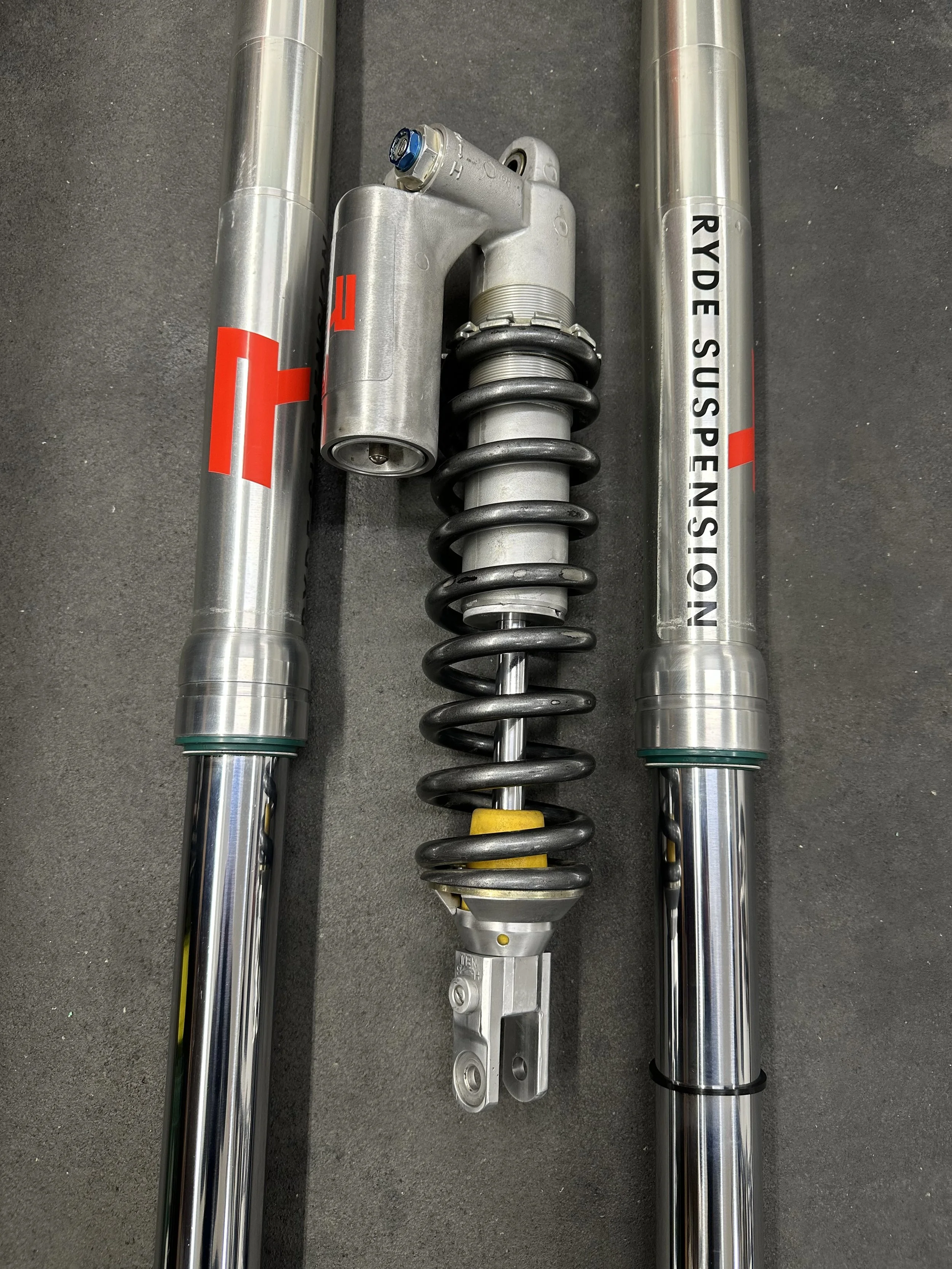 Two silver motorcycle front fork tubes with a black coil spring suspension component in between on a gray surface.