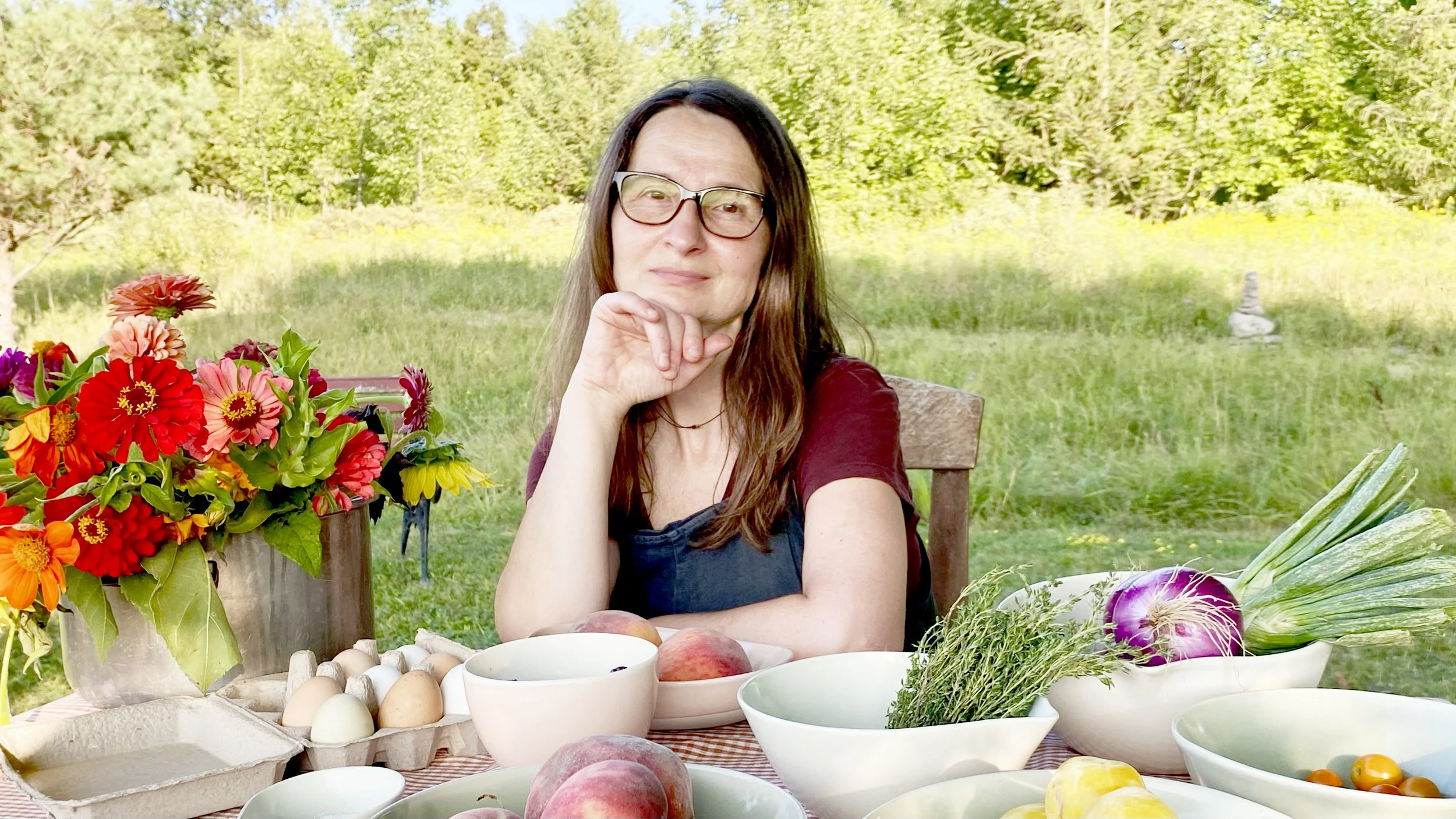 A woman with glasses sitting at a table outdoors with a colorful arrangement of flowers, eggs, peaches, and various vegetables, in a lush green garden.