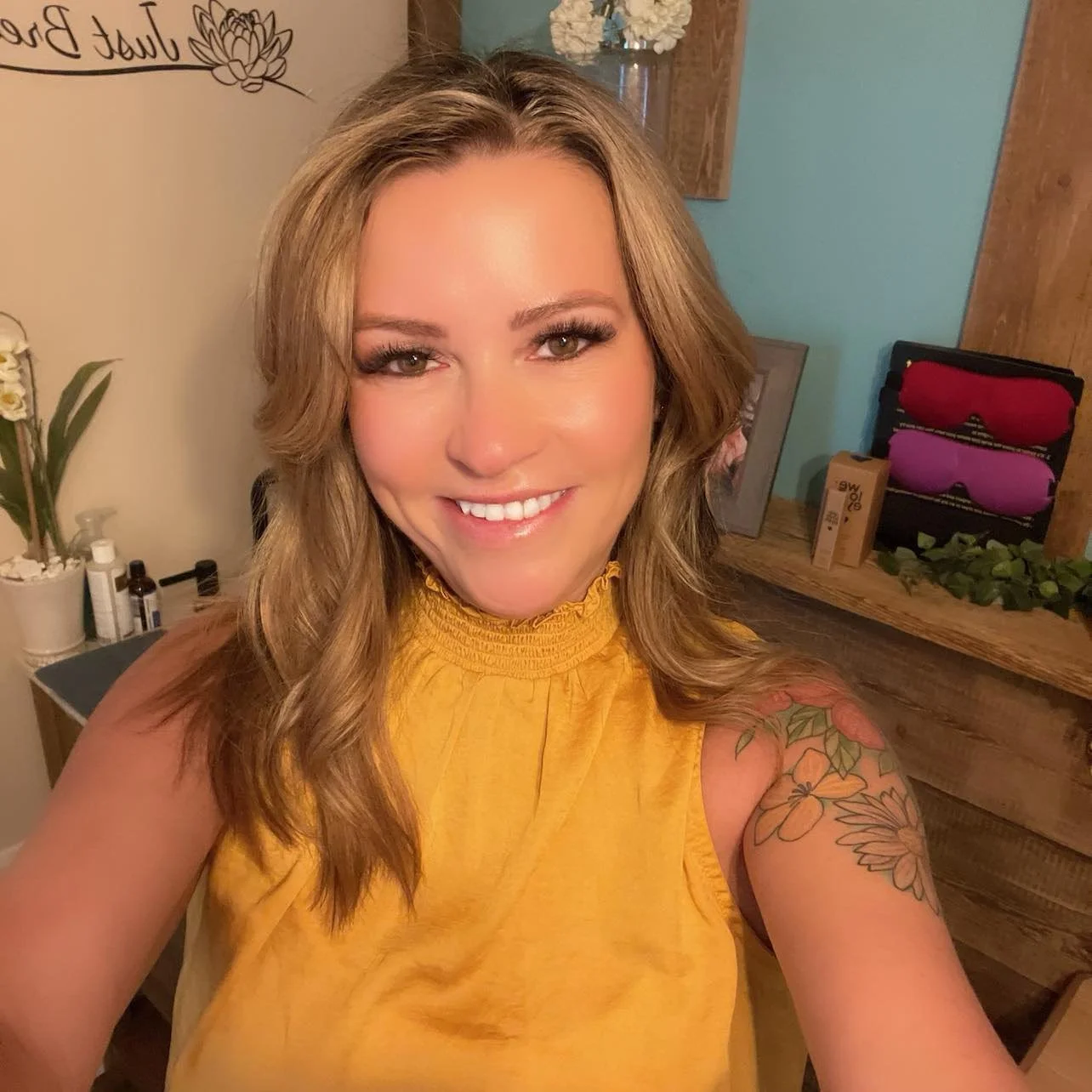 Candice, a smiling woman in a yellow top with a shoulder tattoo, stands indoors surrounded by plants, wooden shelves, and decorative items, reflecting a warm and inviting spa atmosphere.