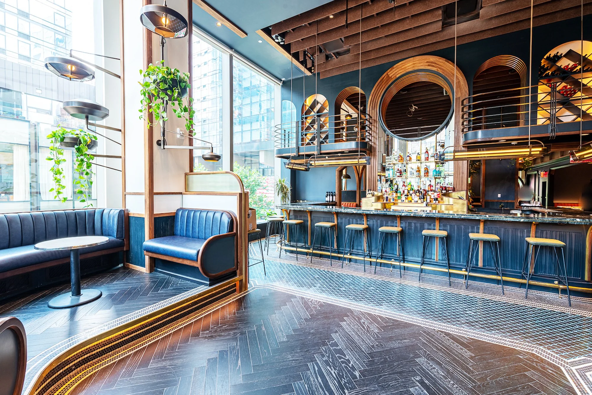 Stylish modern bar with black marble counter, gold accents, and tiered shelving behind with various bottles of liquor. Circular mirror and wooden decorative elements are above the bar, with bar stools lined up in front.