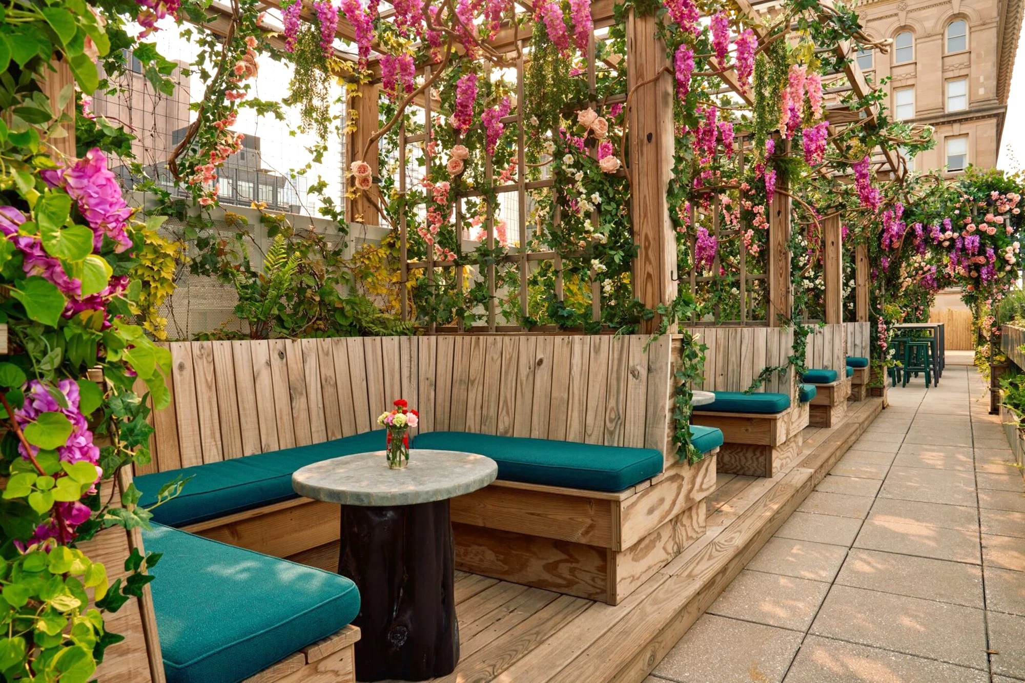Outdoor seating area with wooden benches and turquoise cushions, surrounded by wooden structures and greenery, with a cityscape in the background.
