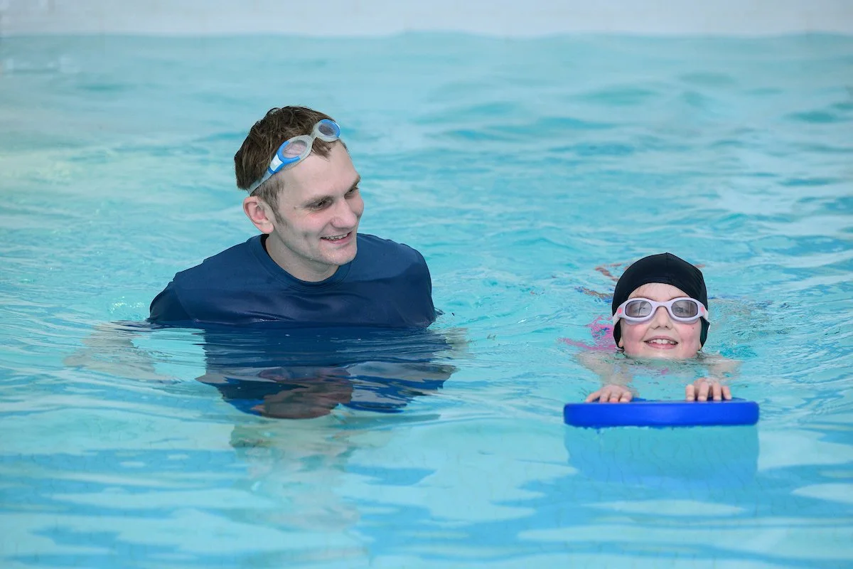 A girl with goggles and a swim cap uses a kickboard while swimming in a pool next to a man wearing goggles on his head. They appear to be having a swimming lesson.