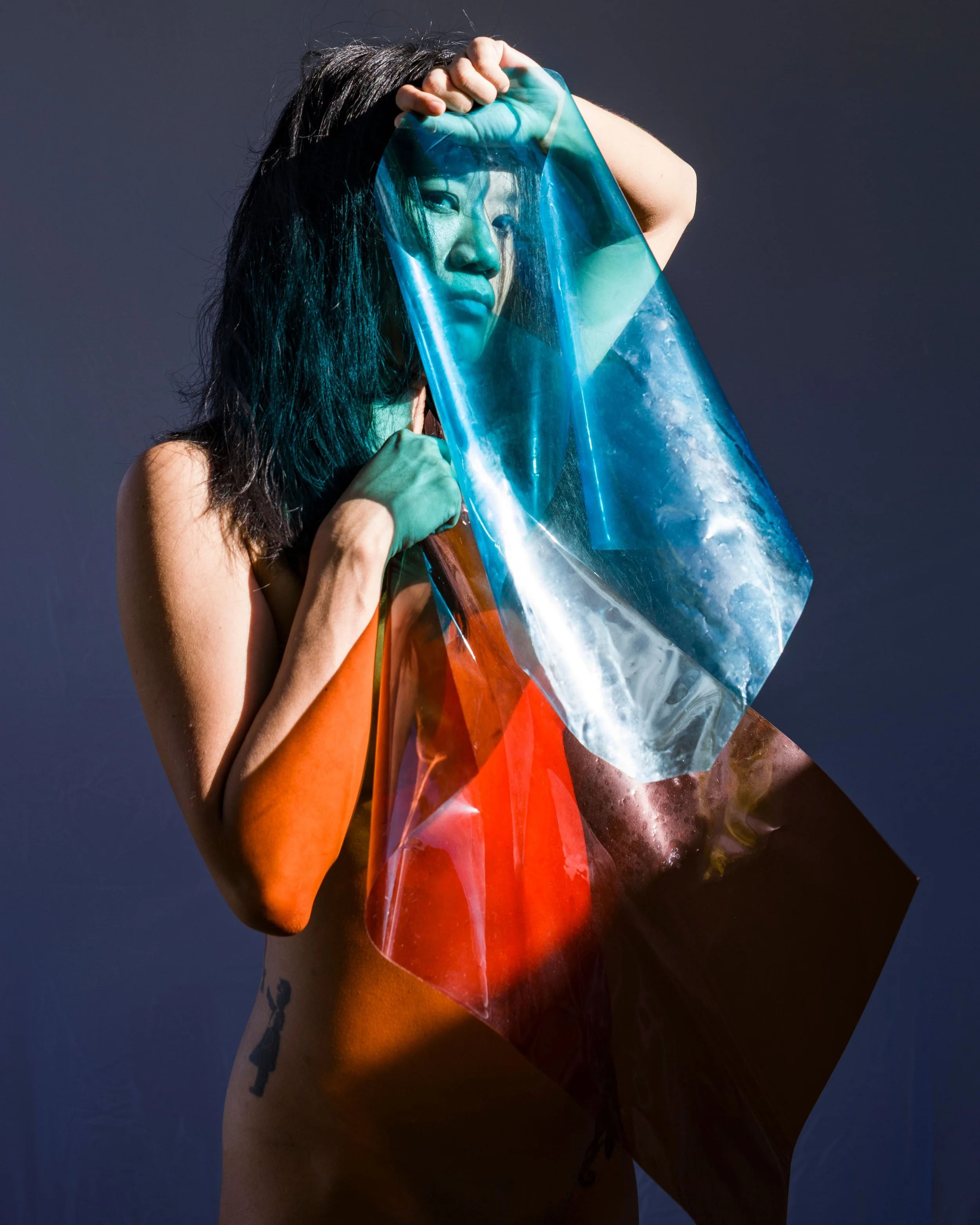 Artist Jenny “Jeta” Tang holds a translucent sheet of blue, red and green cellophane in front of her face against a dark background.