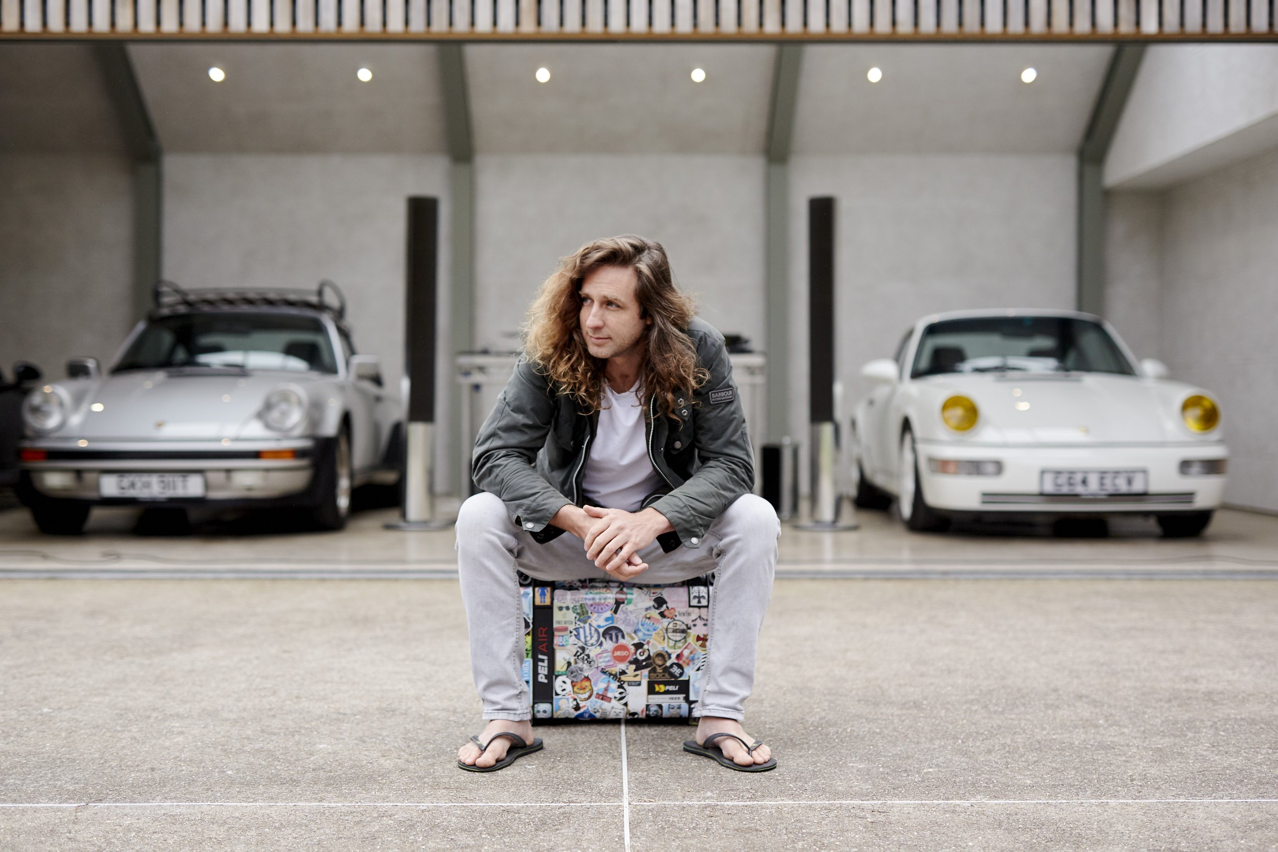 A young man with long, curly hair sits on a luggage case in front of a garage with two white classic cars. He is wearing a dark jacket, white shirt, light pants, and flip-flops, gazing to the side.