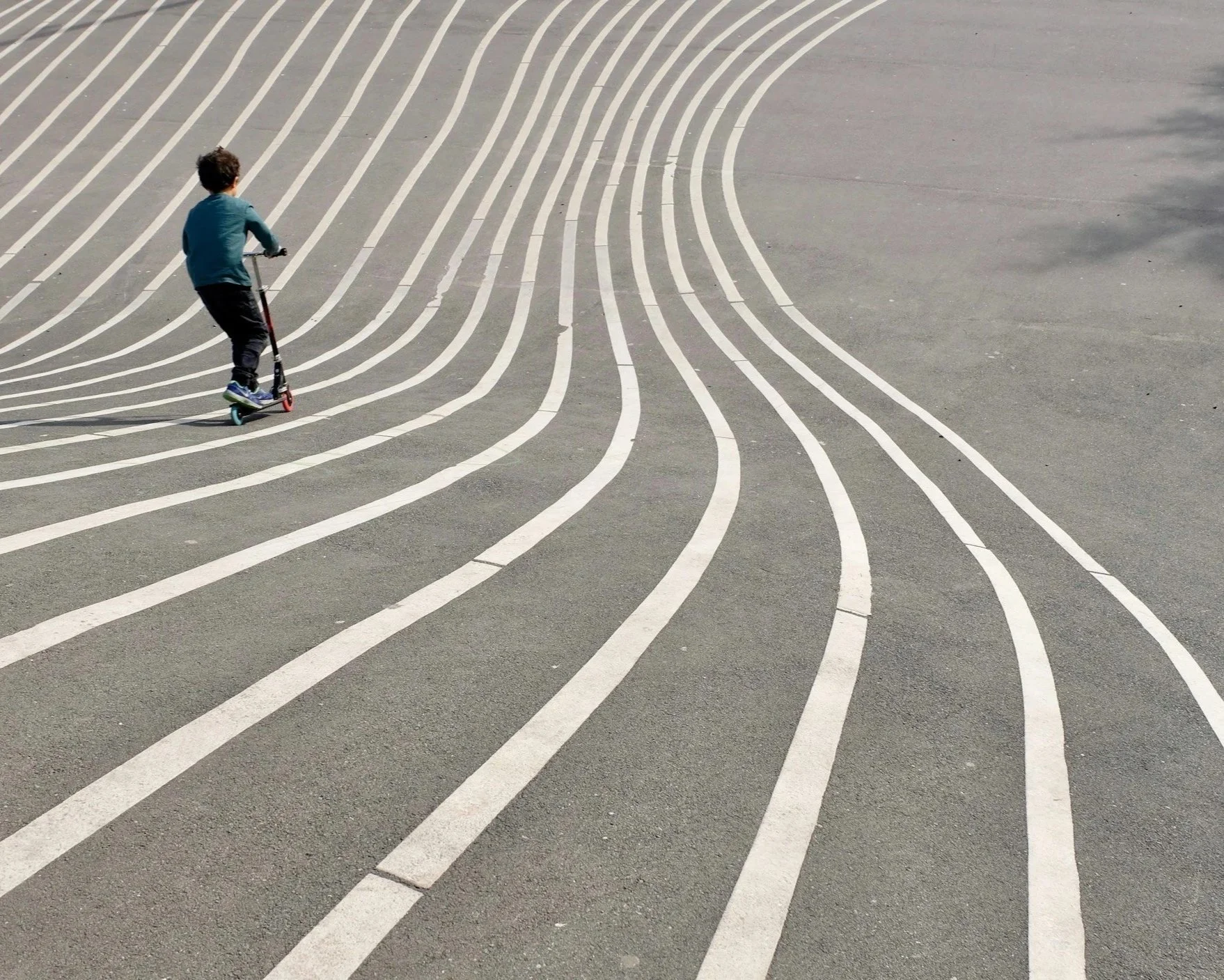 Child riding a scooter along curved white lane markings on a paved surface, viewed from above.