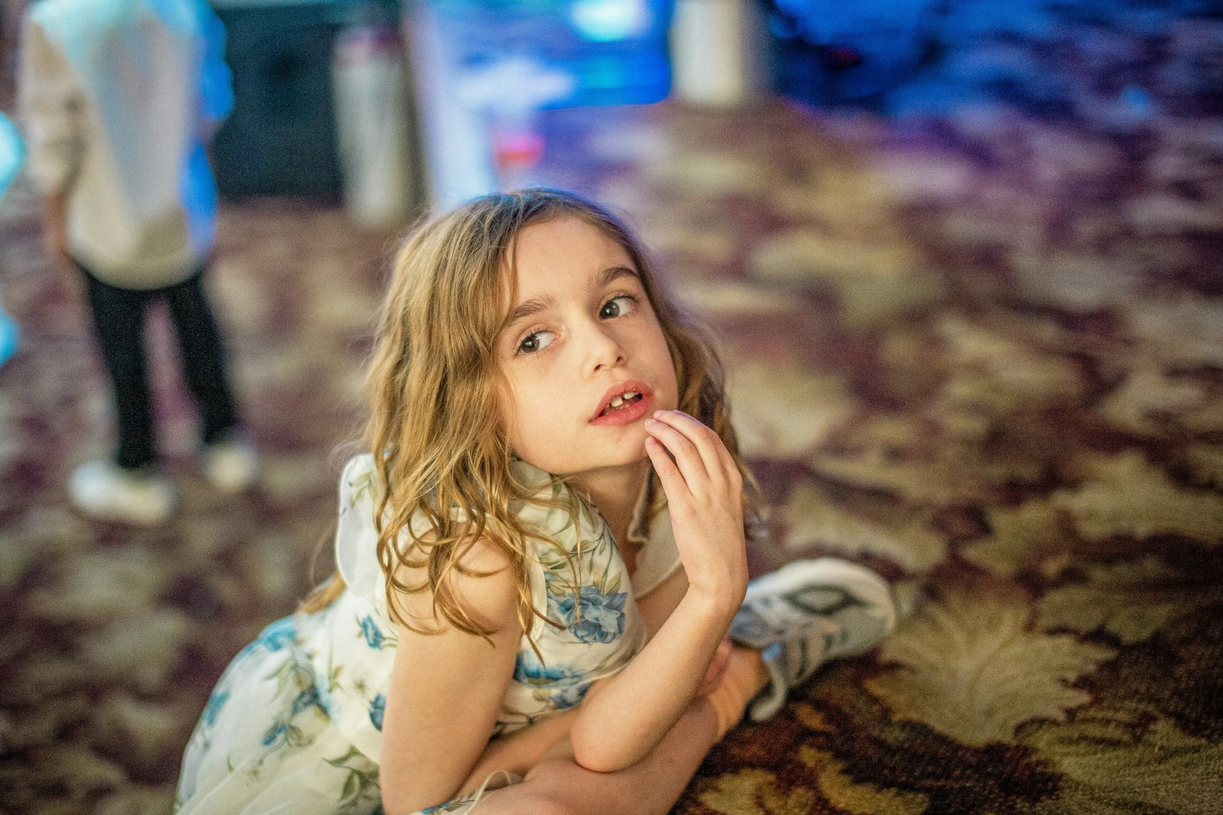 Young girl sitting on the floor and looking upward while adults stand blurred in the background.