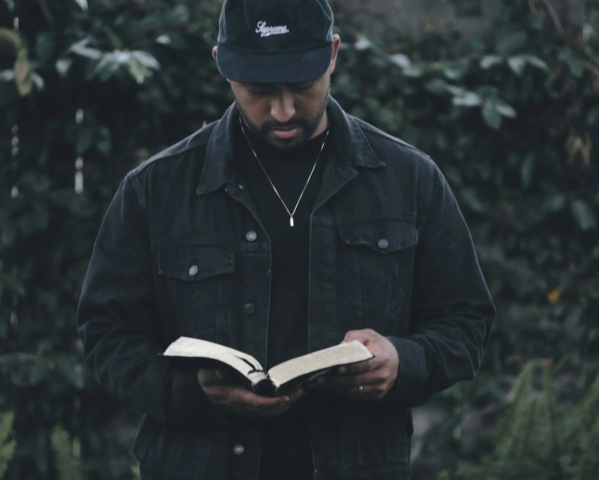 Man reading a religious book outdoors with green bushes in the background symbolising spirituality and reflection.