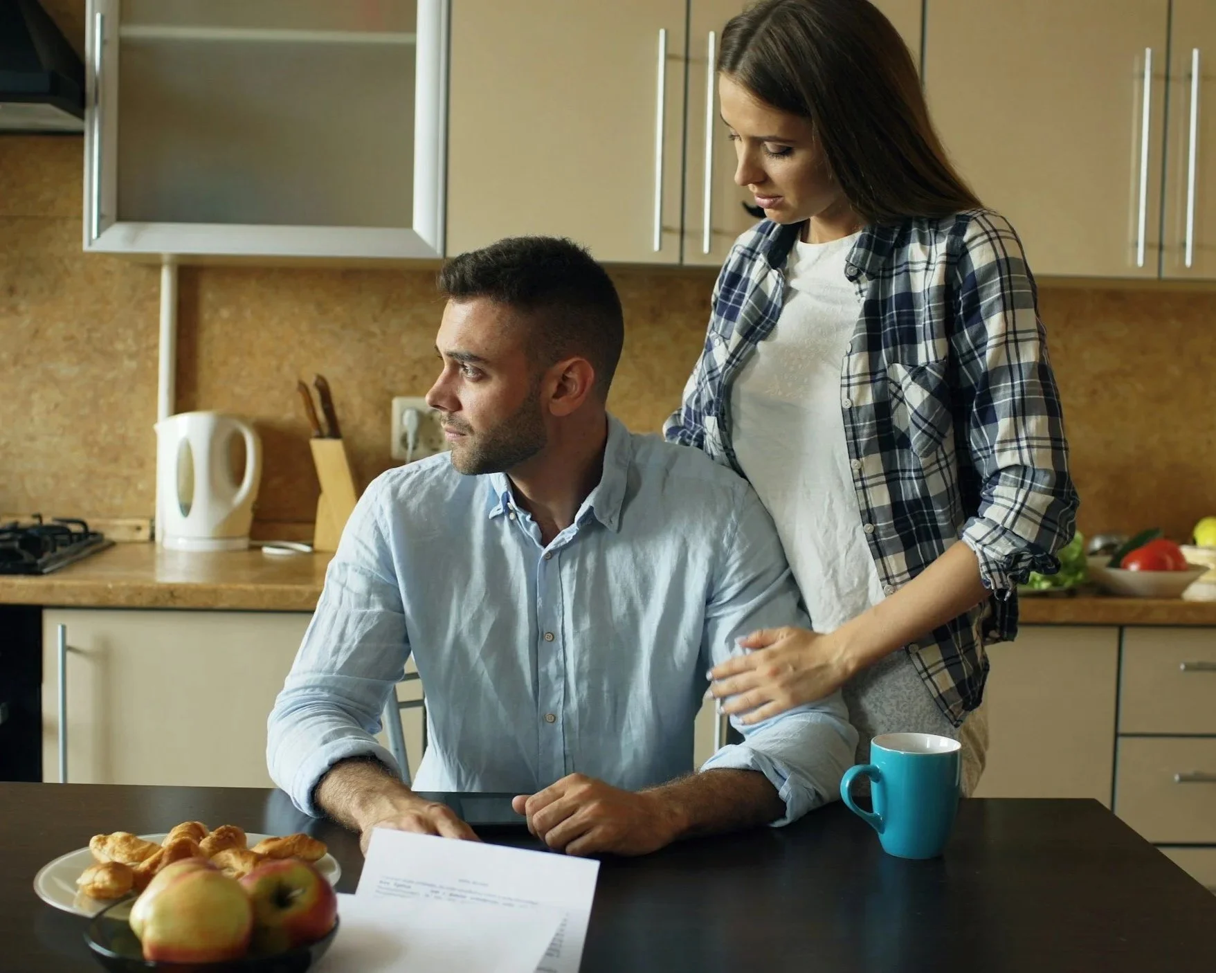 Person sitting at a kitchen table looking worried while a woman stands behind them, illustrating emotional responsibility within a relationship.