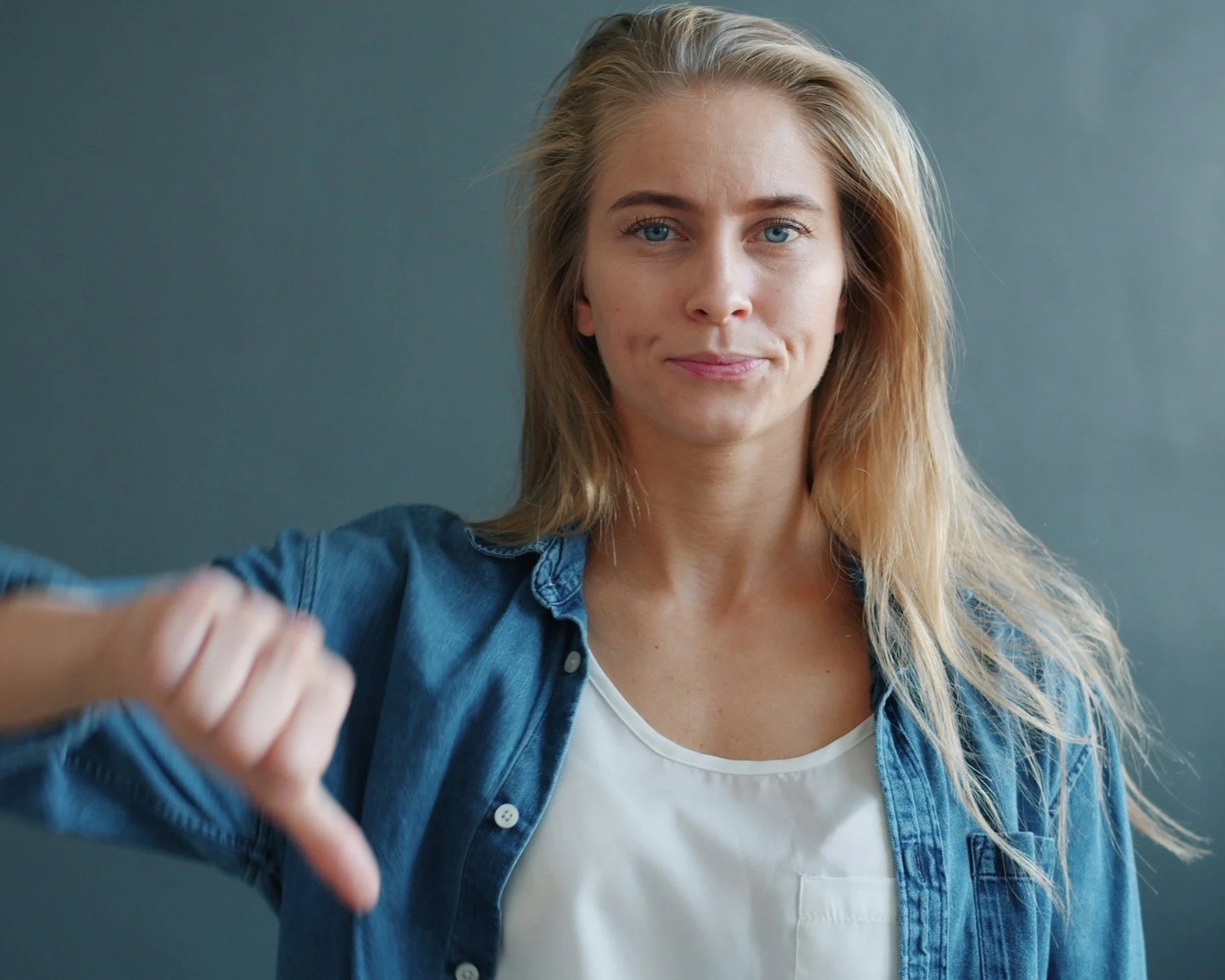 Young blonde woman in a denim shirt giving a thumbs-down gesture, representing self-criticism, disapproval and the internalised voice of the inner critic.