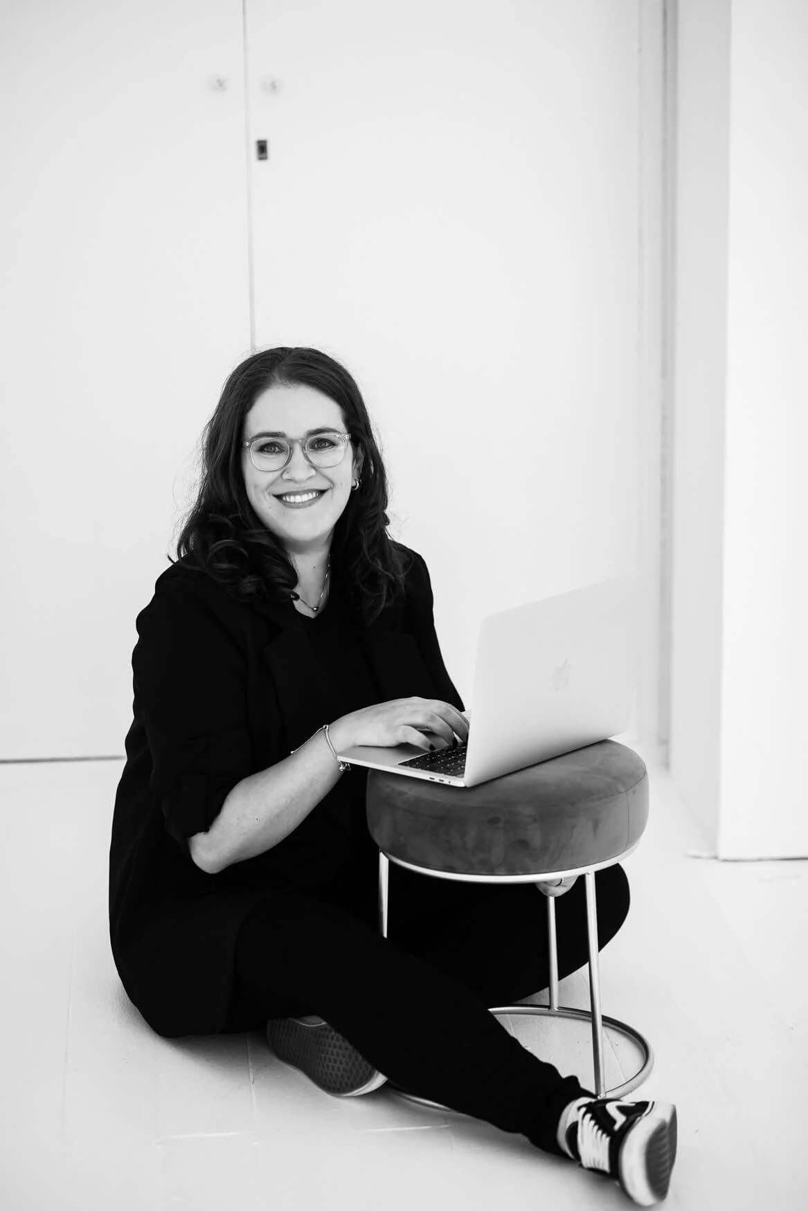 A woman with glasses and dark hair sitting on the floor, smiling, working on a laptop placed on a small round stool.