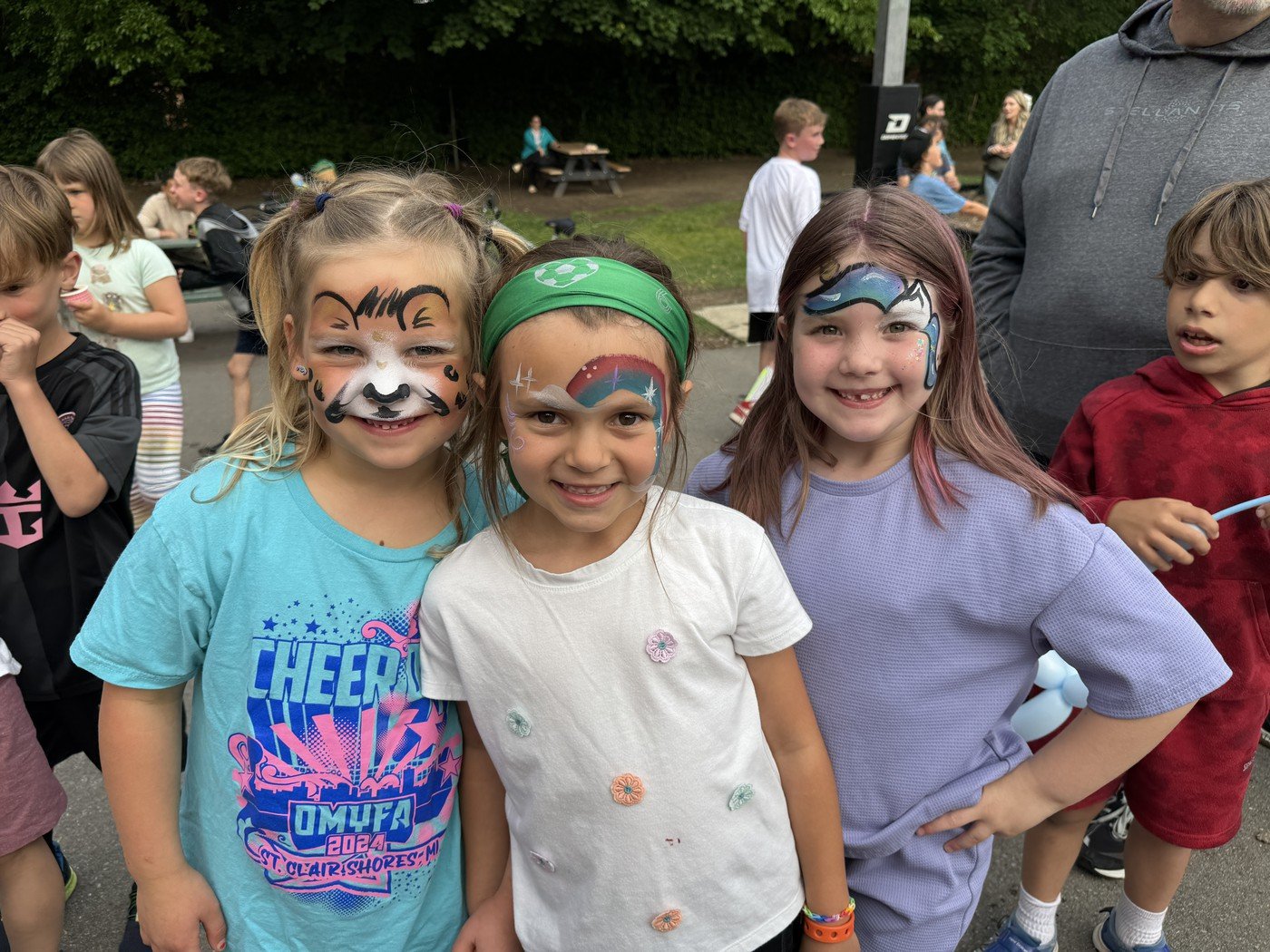 Three young girls with face paint, smiling at the camera at an outdoor gathering, with other children and adults in the background.