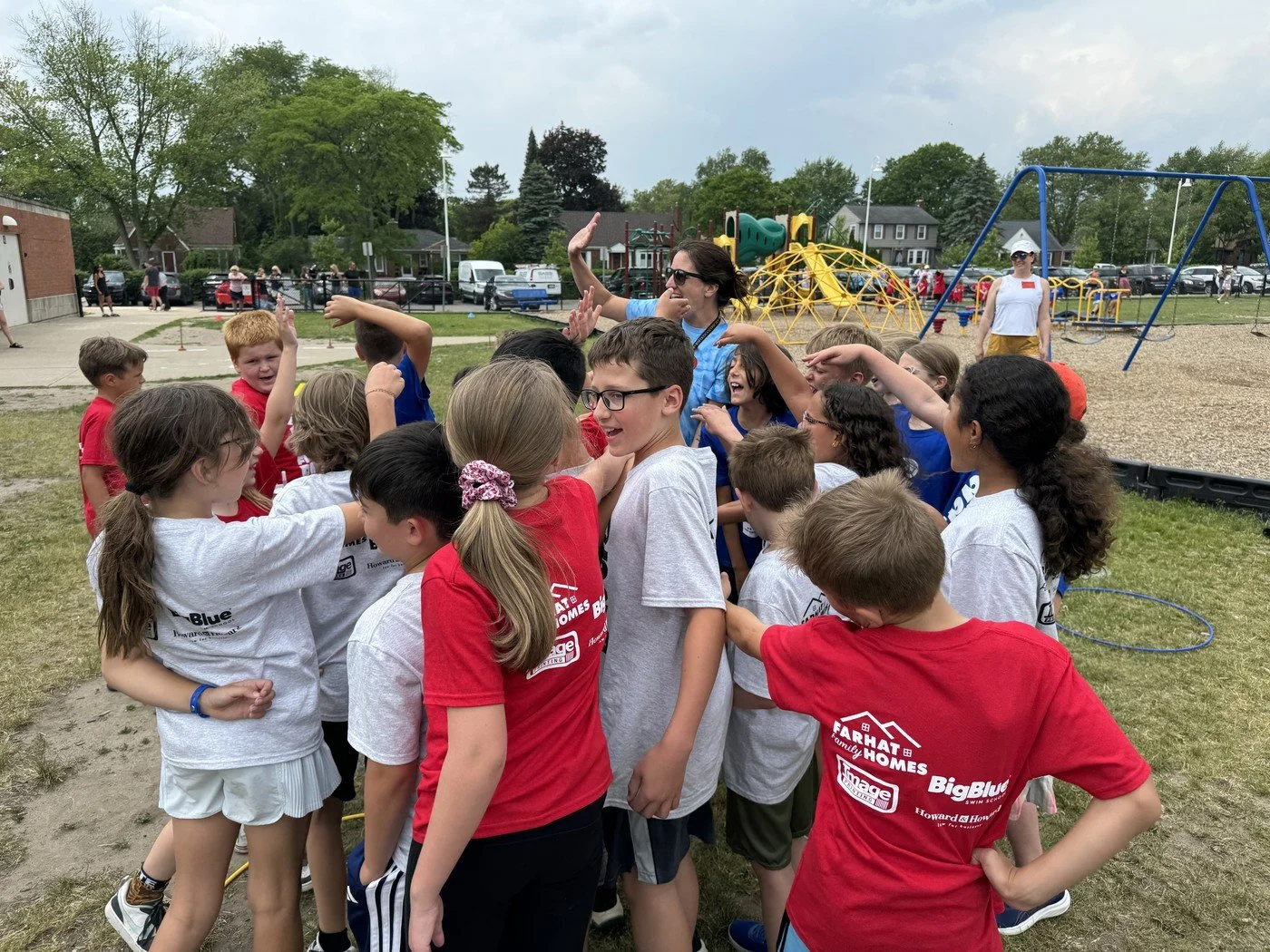 A group of children and a woman on a playground. The children are wearing colorful t-shirts and are engaged in a group activity or game, with some smiling and raising their hands. The woman is wearing sunglasses and a blue shirt, participating in the