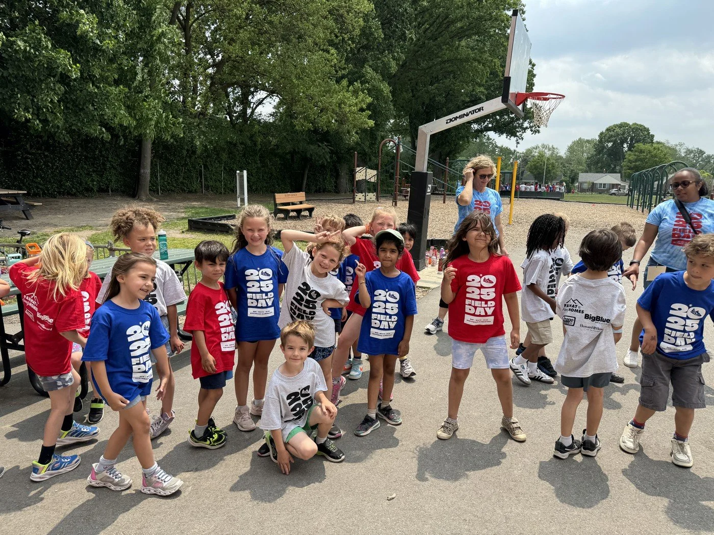 Children gathered outdoors on a paved area for a school or community event, with some wearing red, white, or blue t-shirts that say 'FIELD DAY 2023,' and adults supervising in the background near a basketball hoop and a playground.