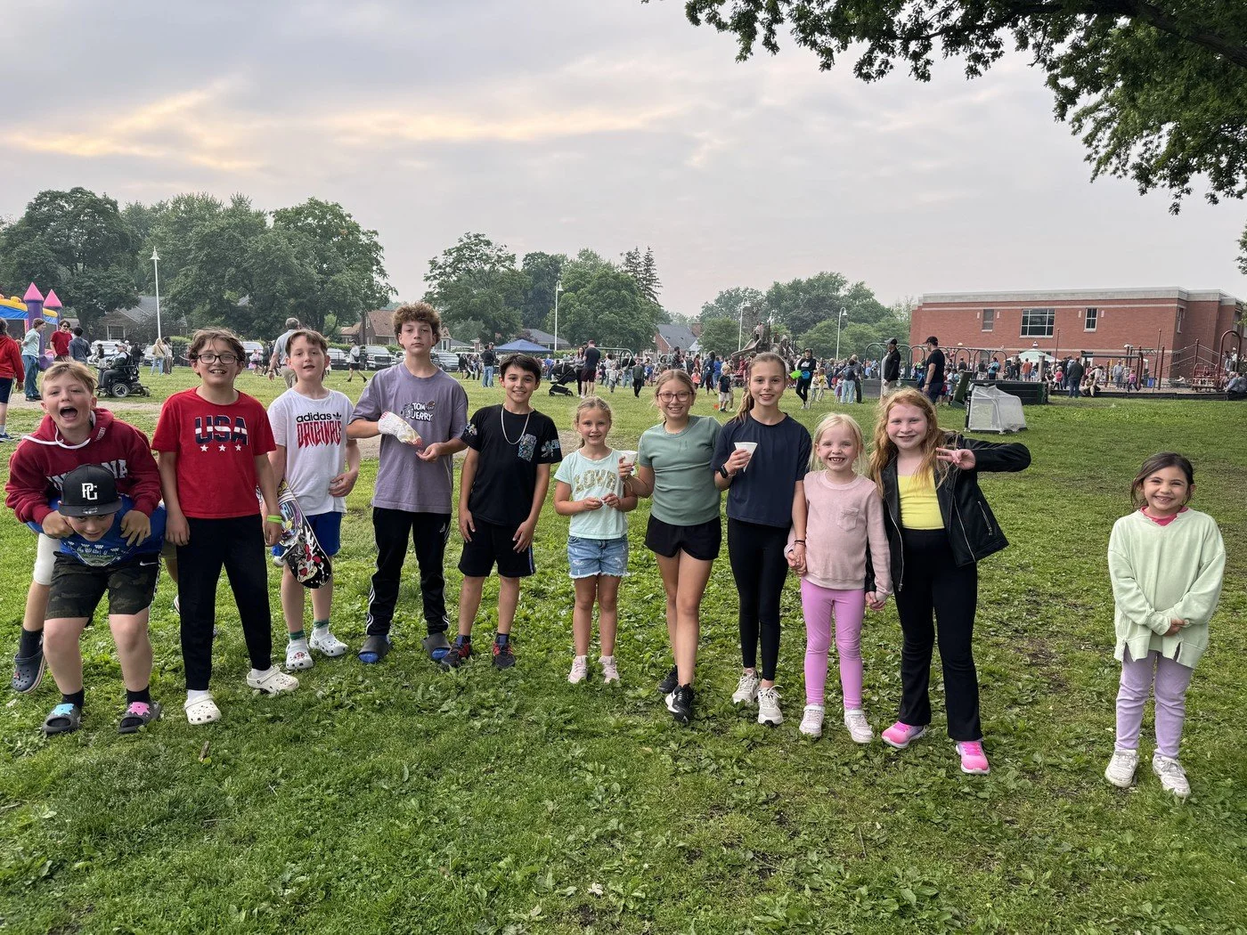 Group of children standing on grass at an outdoor event or park, with other people and a building in the background.