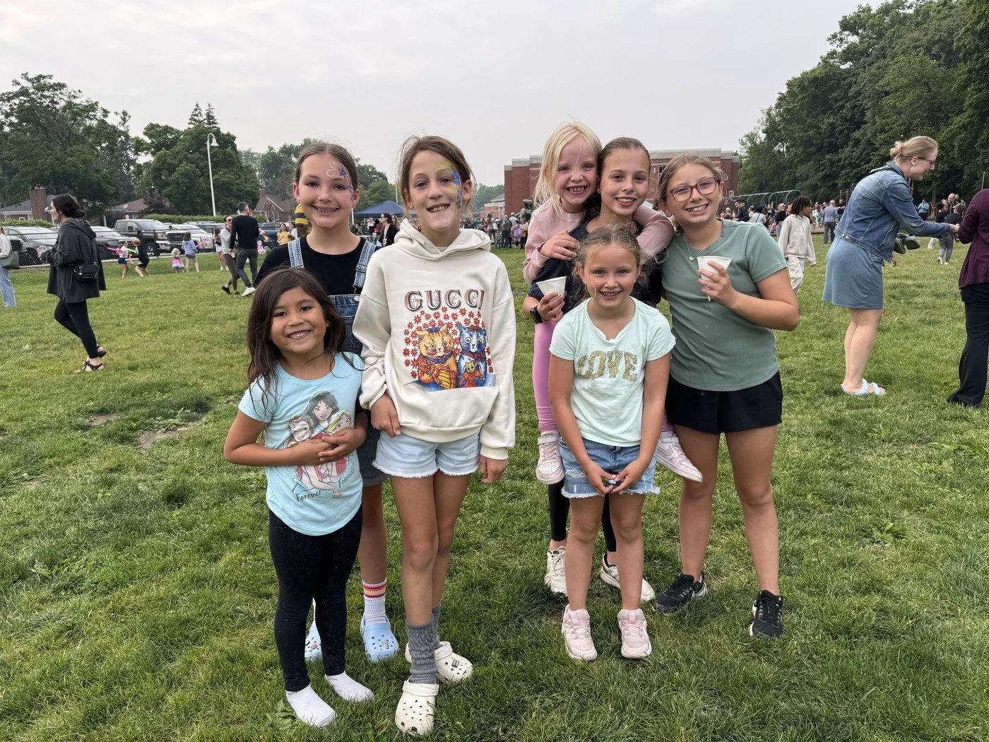 Group of eight girls standing on grass at an outdoor event, smiling and posing for the camera, with more people in the background.