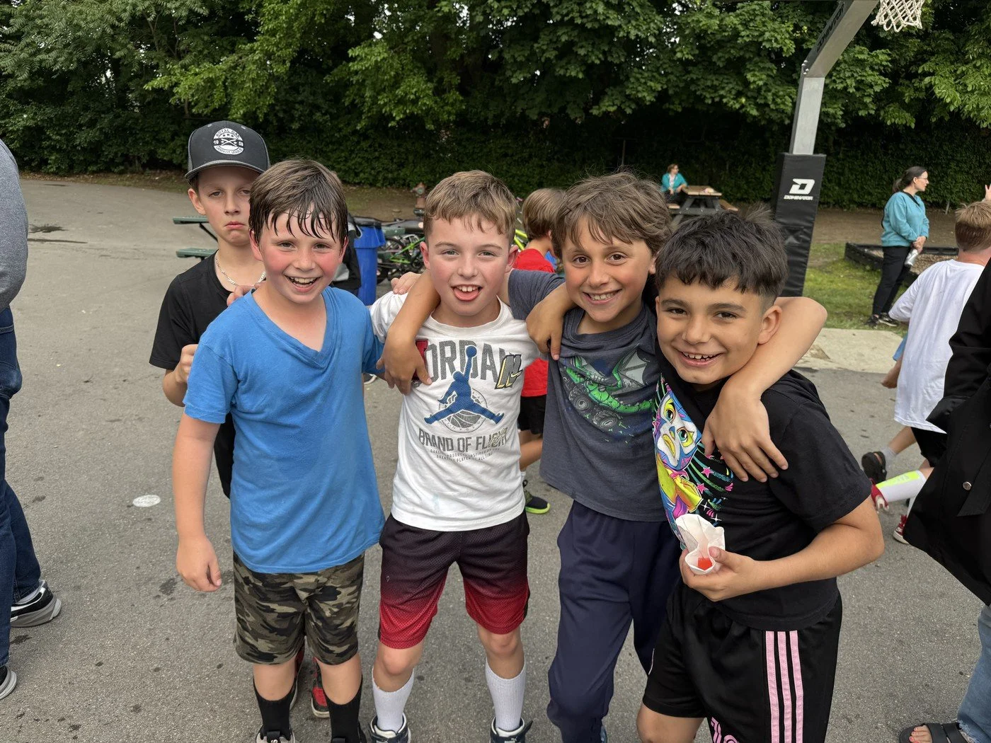 Group of five young boys smiling and hugging at a school outdoor event, with basketball court and trees in the background.