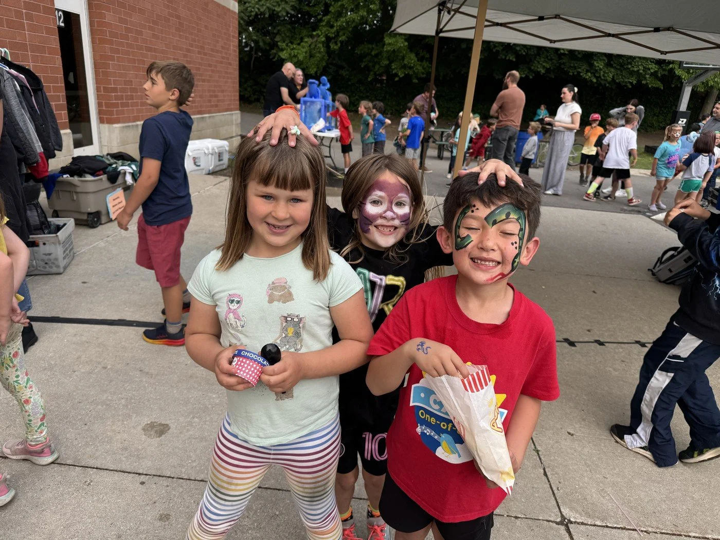 Three children smiling and posing with face paint and face masks at an outdoor event, possibly a fair or festival, with other kids and adults in the background.