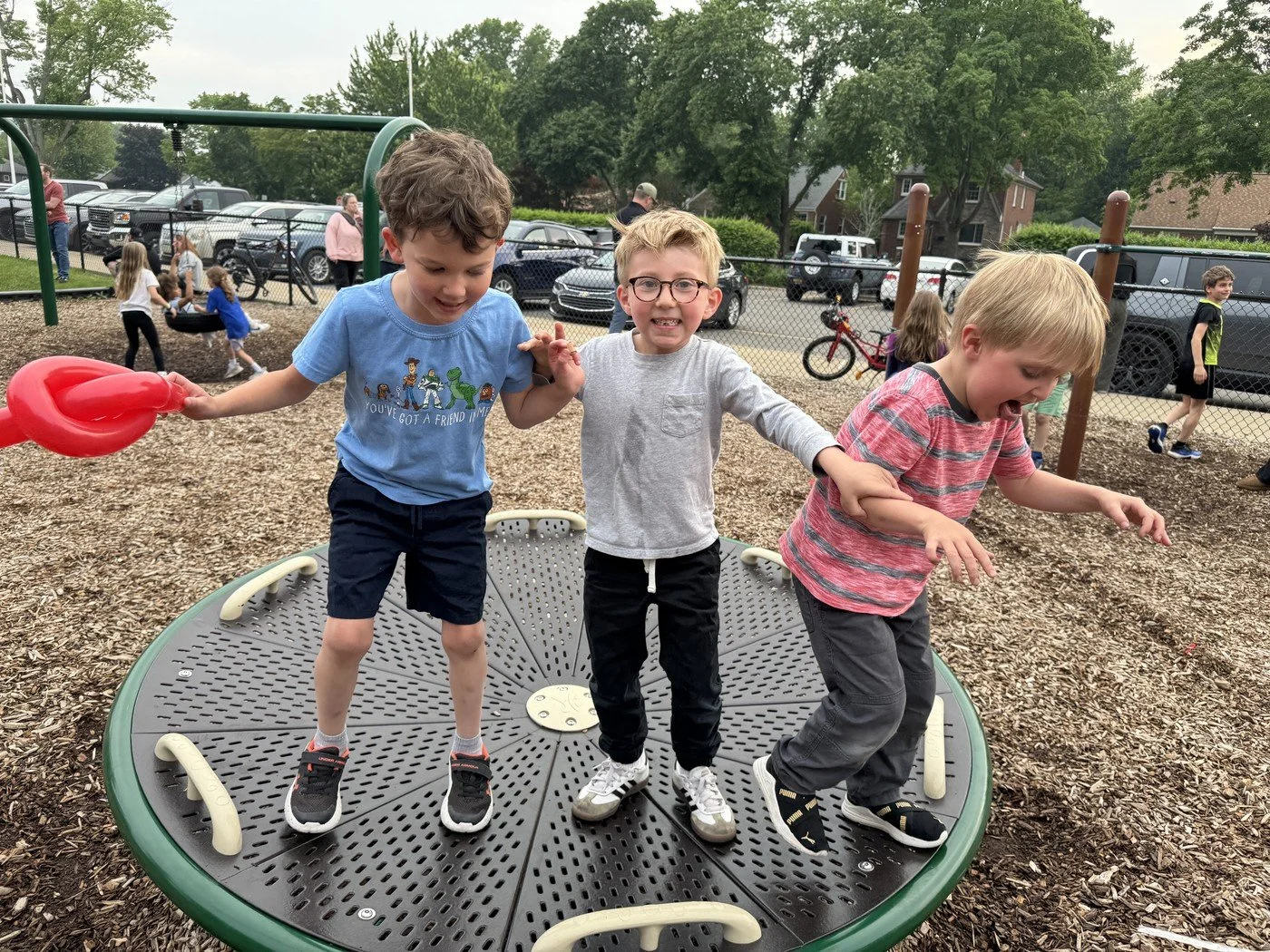 Three young boys holding hands and jumping on a circular playground spinner at a park on a cloudy day.