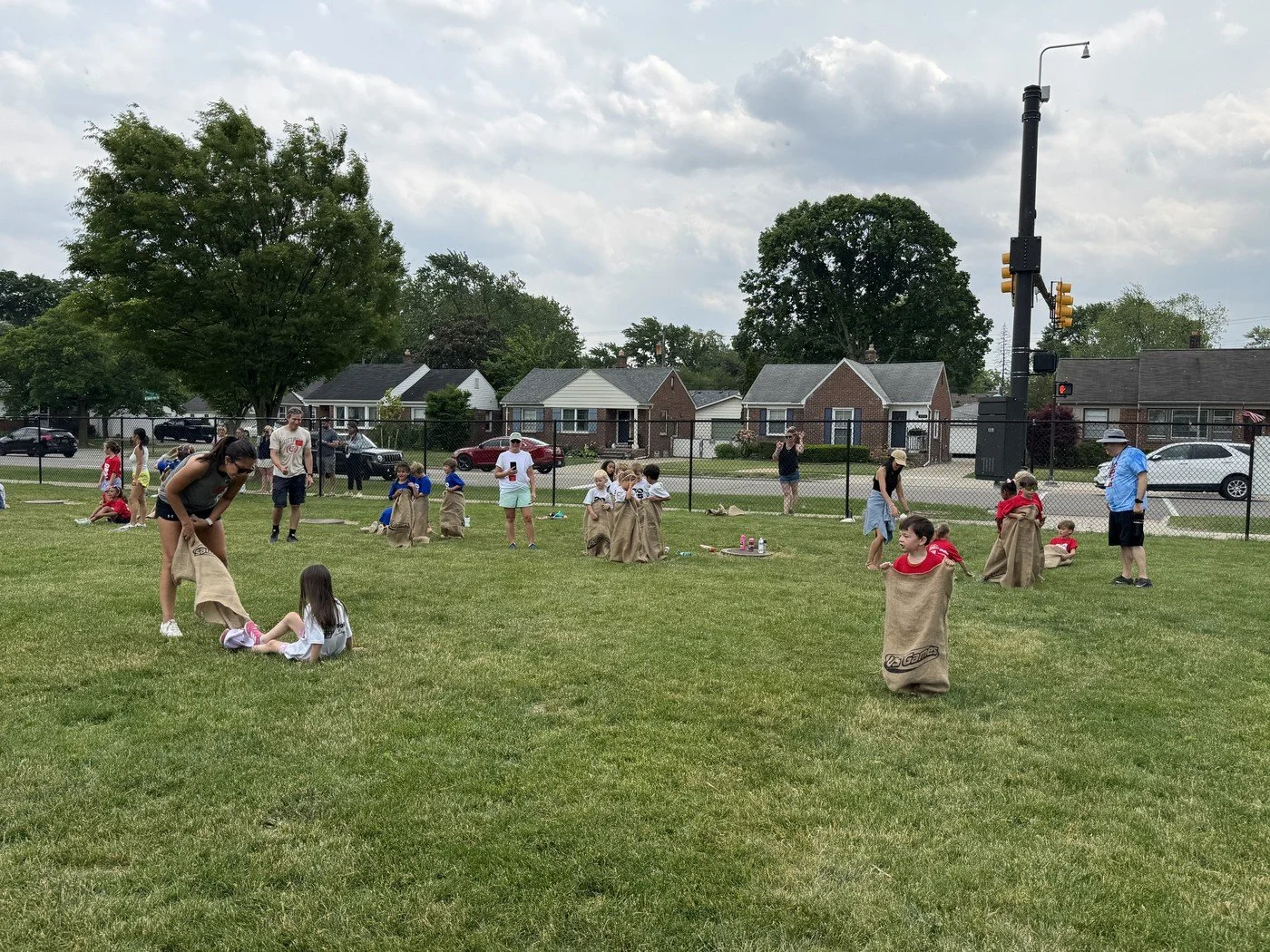 Children and adults participate in a sack race on a grassy field, with houses and trees in the background, under a partly cloudy sky.