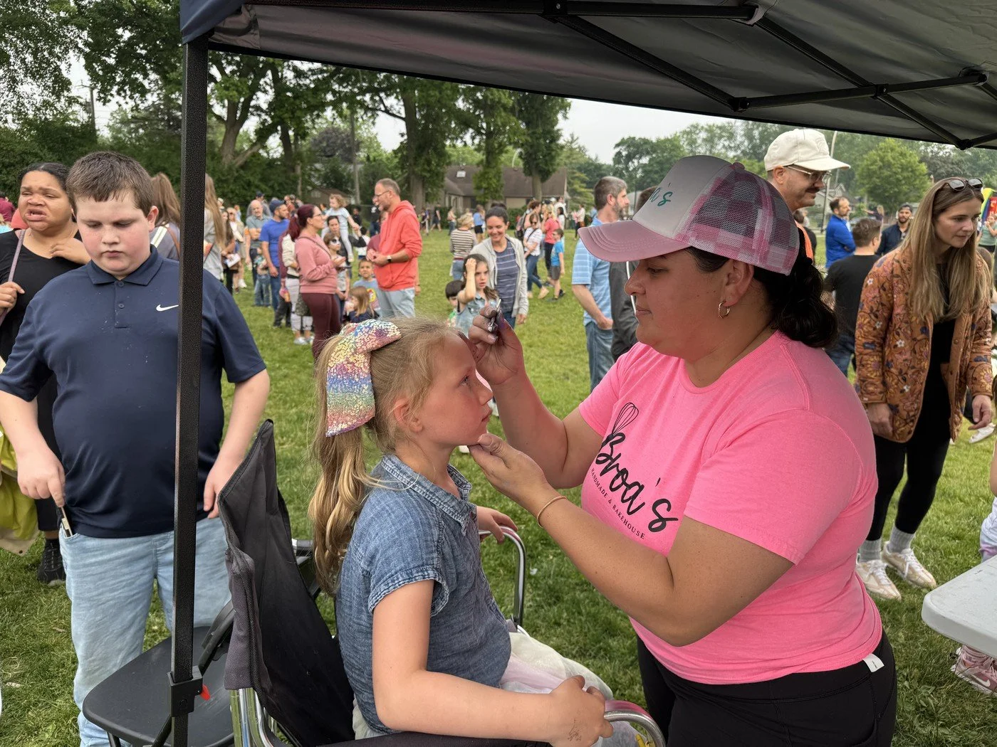 A woman wearing a pink T-shirt and pink baseball cap is applying face paint to a young girl sitting in a wheelchair at an outdoor event. There are many people and children in the background on a grassy field.