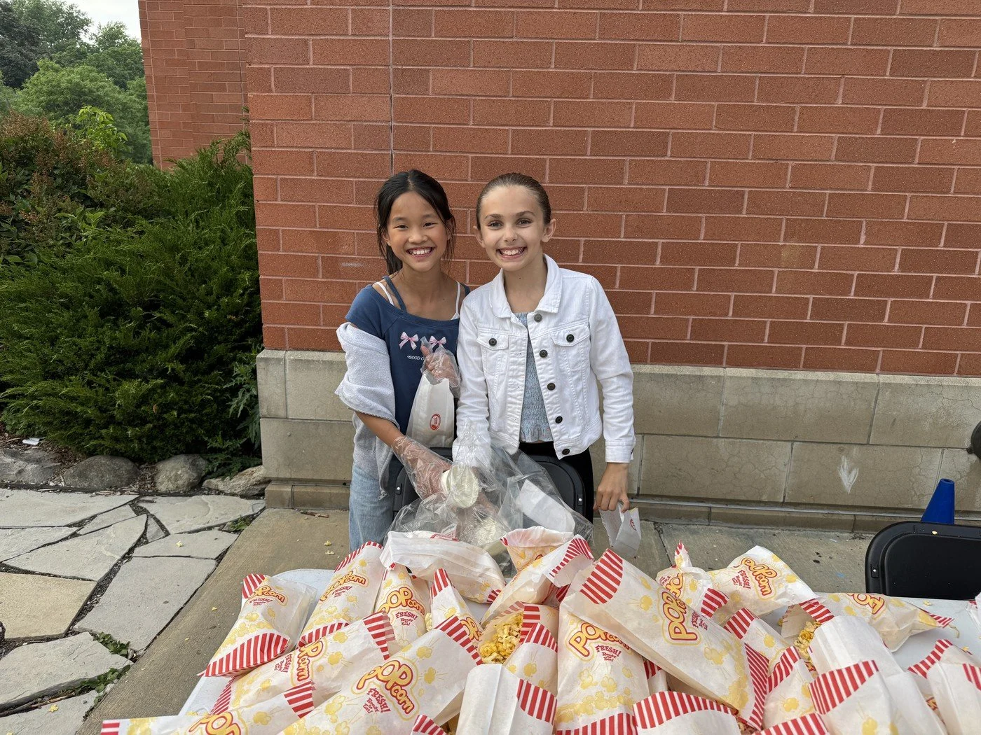 Two smiling girls standing behind a table piled with many bags of popcorn outdoors, with a brick wall and greenery in the background.