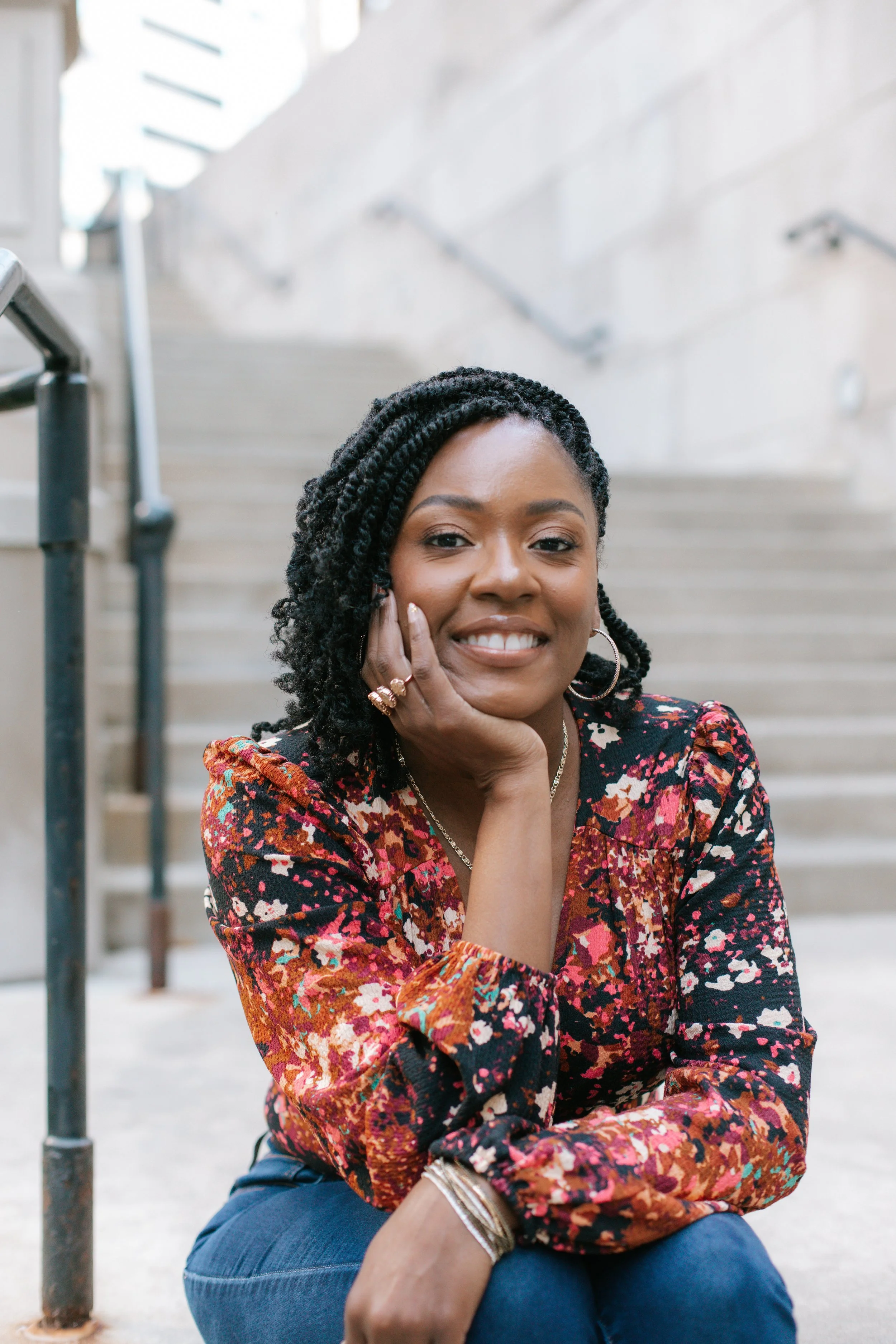 Woman with curly hair in a floral long-sleeve top sitting outdoors on stairs, smiling with her chin resting on her hand.