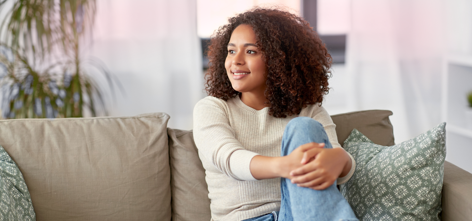 A woman with curly brown hair smiling while sitting on a beige sofa with patterned pillows in a bright living room.