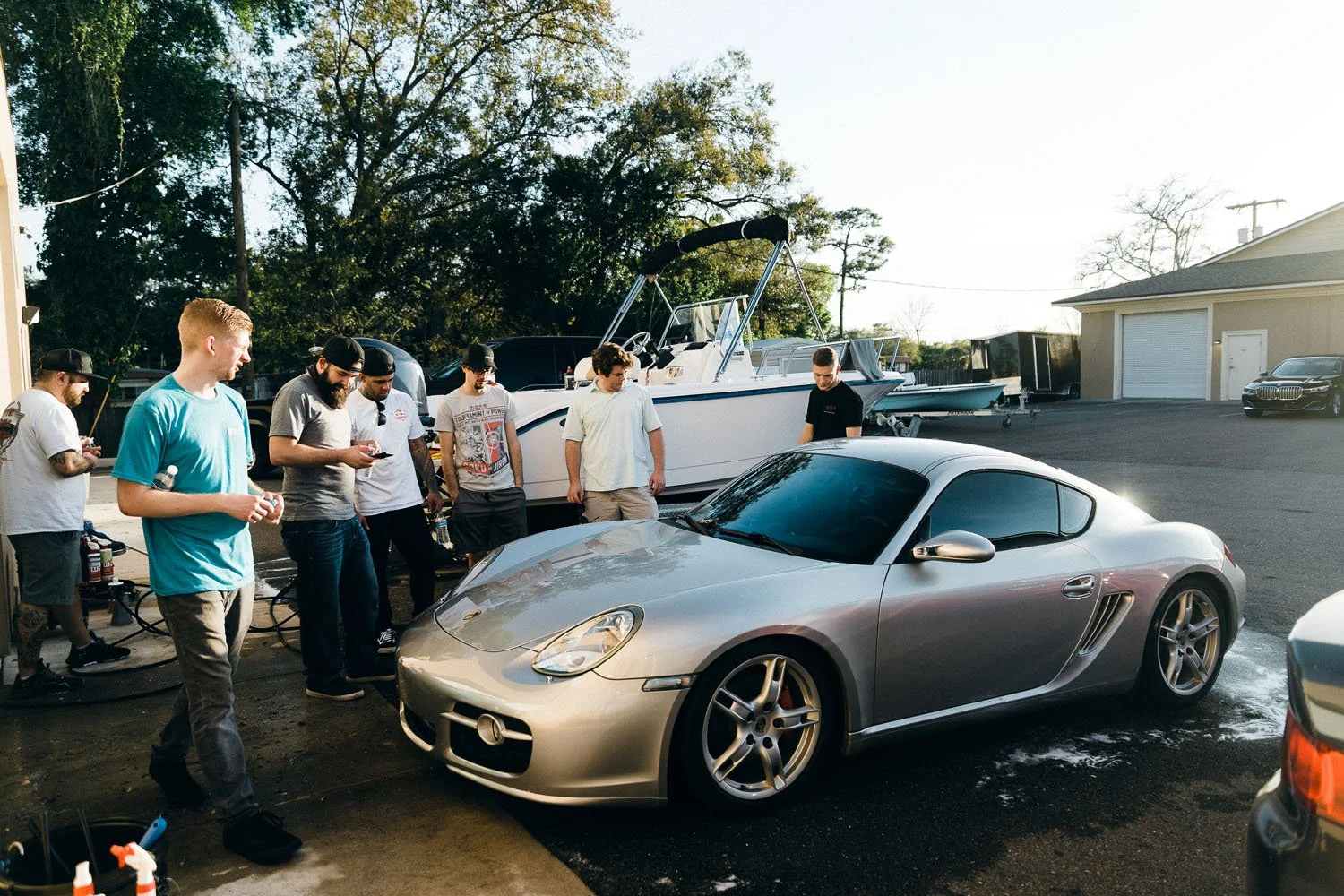 A group of men washing a silver sports car in an outdoor car wash area with a large boat on a trailer and trees in the background.