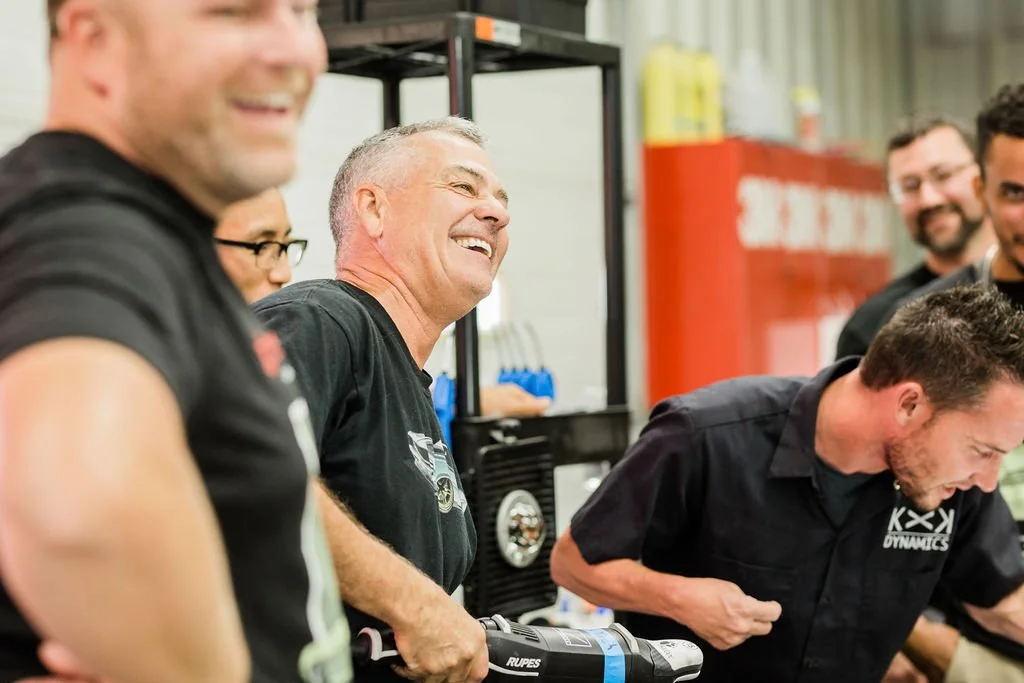 Men sharing a laugh and talking in a workshop setting, with machinery and tools in the background.
