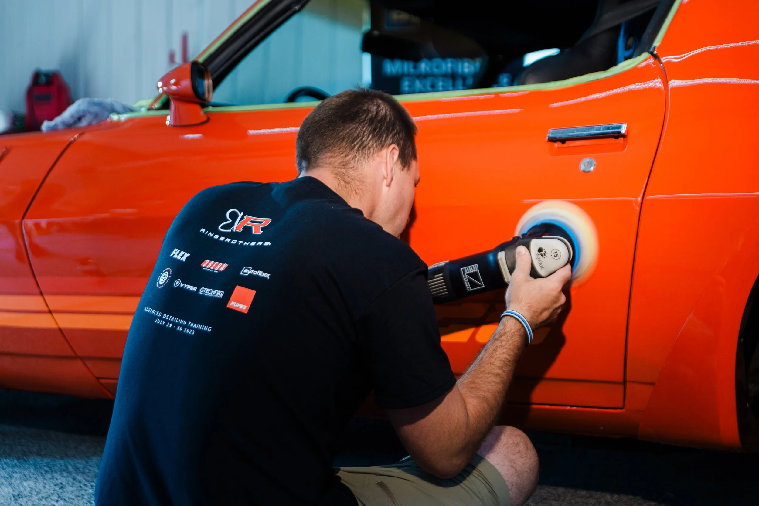 A person polishing an orange car with a rotary buffer in a garage or workshop.