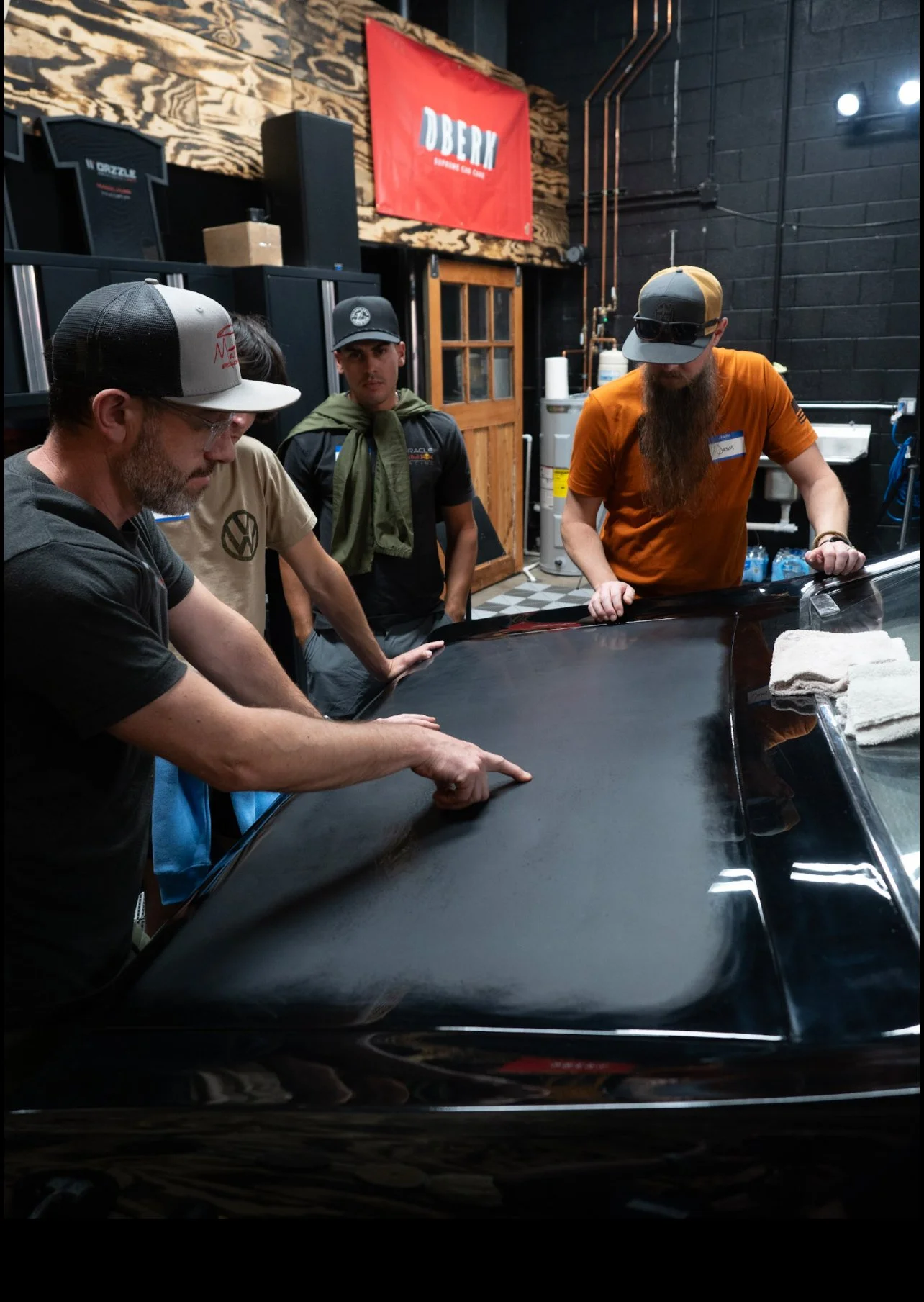 Four men examining the hood of a black car in a workshop or garage, with a red banner that says 'DUBN' hanging on the wall behind them.