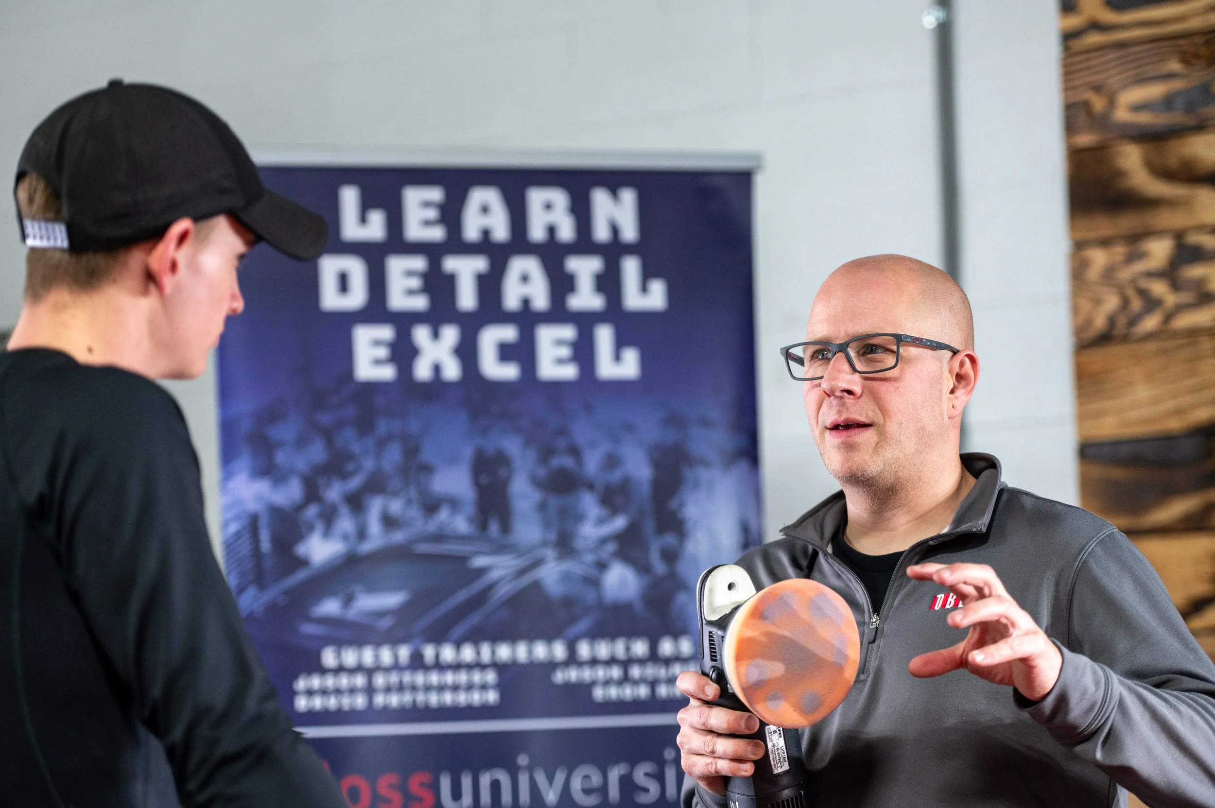 Two men having a conversation in a gym. One man is wearing a black cap, and the other is bald with glasses. Behind them, there is a poster that says 'Learn Detail Excel' with a group of people in the background.
