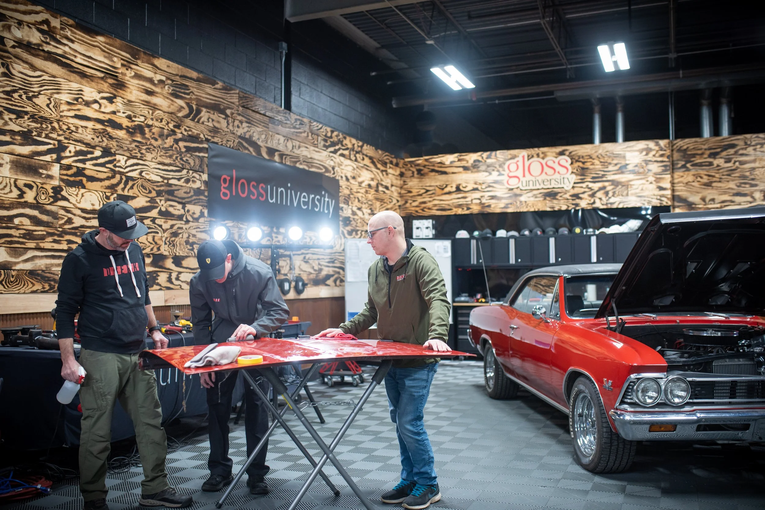 Three men working on a red car inside a garage at Gloss University, with a wooden wall background and a sign that says 'gloss university'. The car's hood is open, and the men are examining or working on the vehicle. There are tools and equipment on a table nearby.