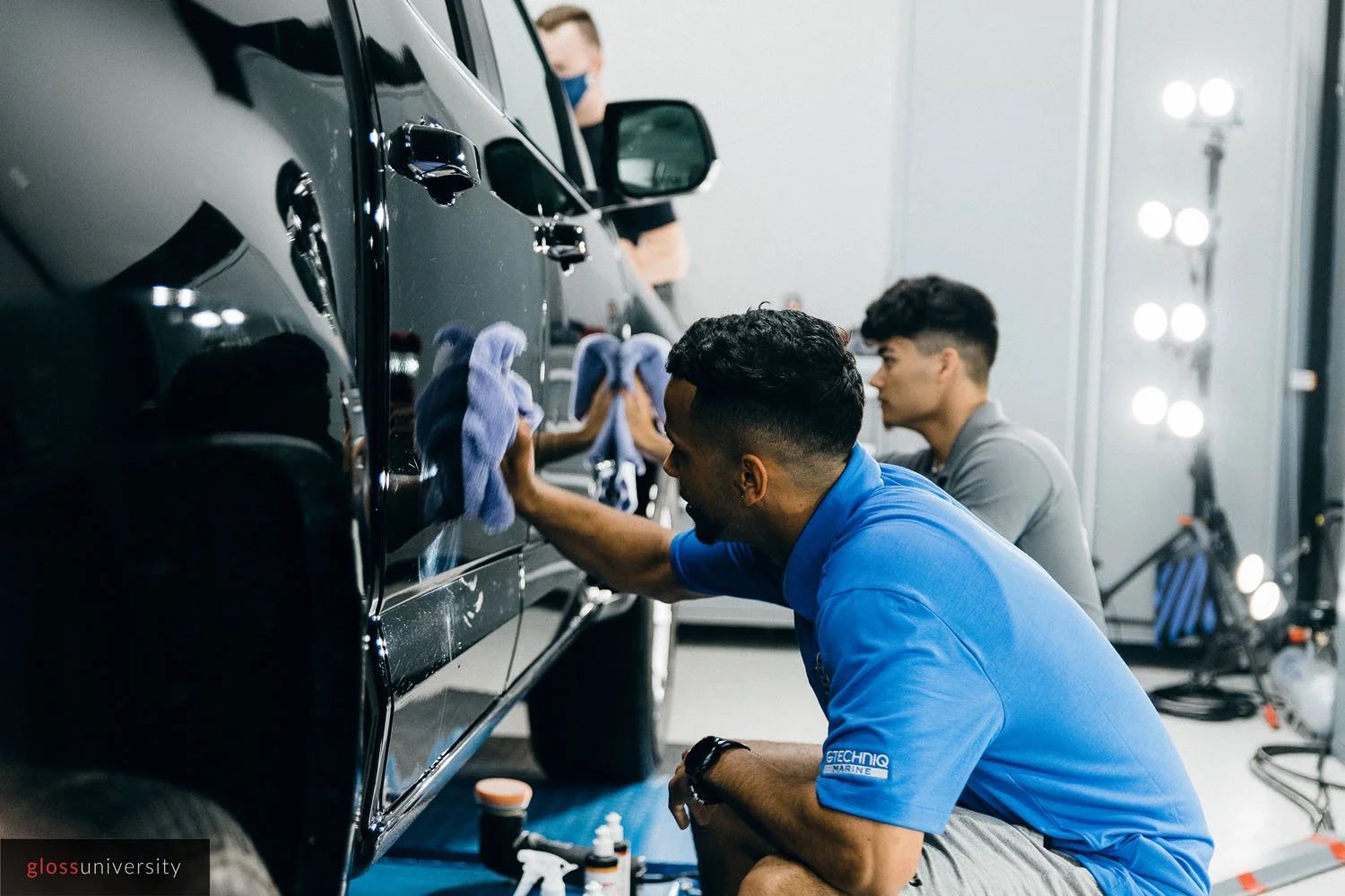 Two men detailing a black car, one kneeling and cleaning the car with a cloth, and the other sitting behind him observing. Both are indoors in a well-lit garage or studio.