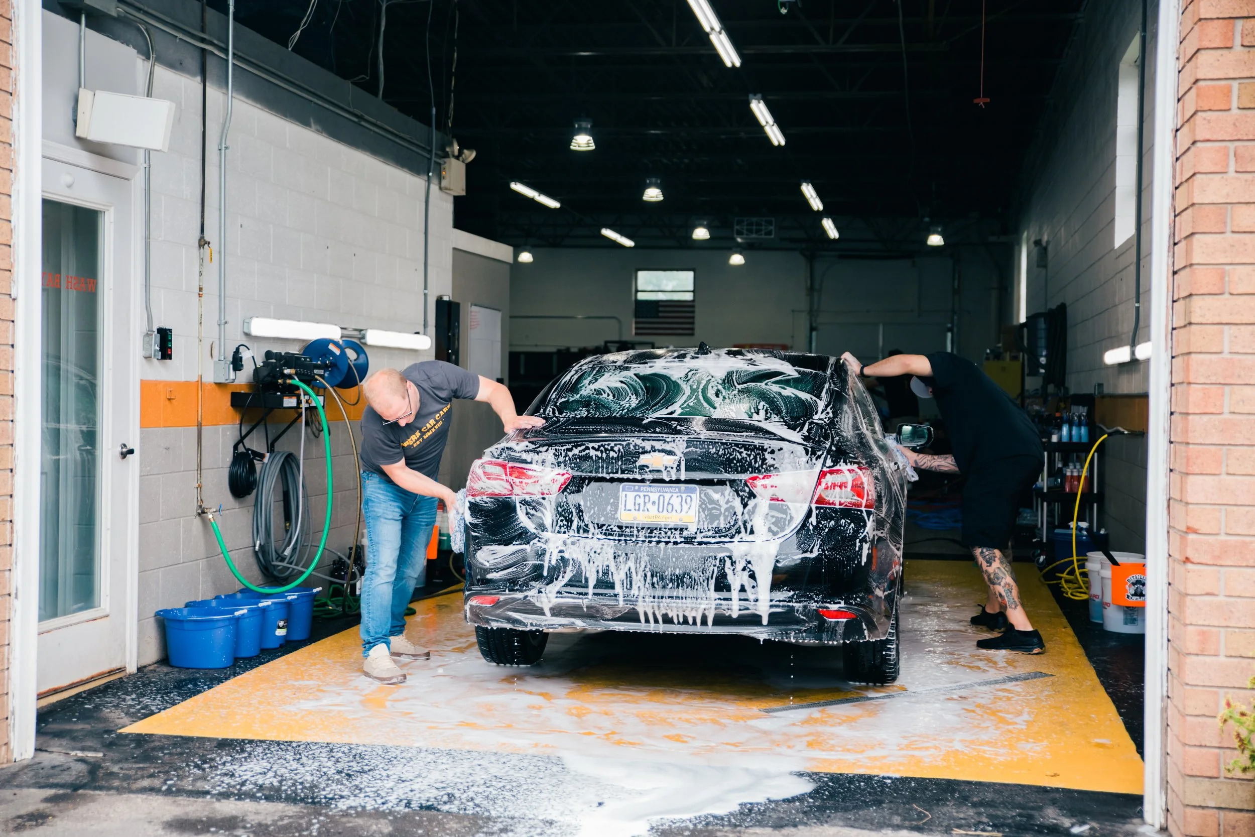 Two people wash a black sedan car with soap inside a garage.