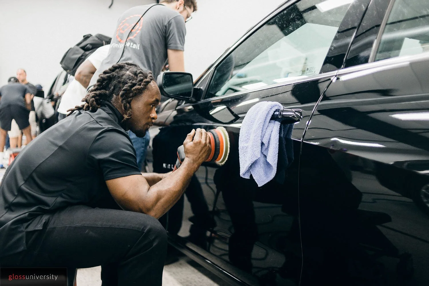 A man with dreadlocks polishing a black car with a polishing tool, a towel hangs from the car's side mirror, and several other people are in the background in a workshop or garage setting.