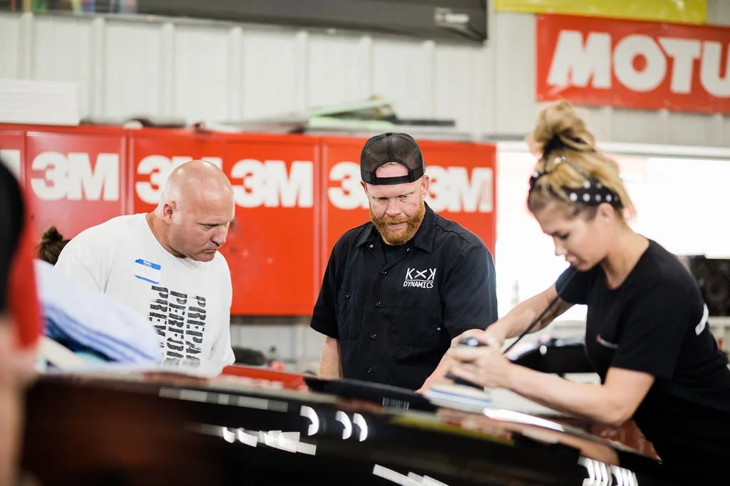 Three people working together in an auto workshop, looking at documents or a laptop, with car parts and 3M and Motul signs in the background.