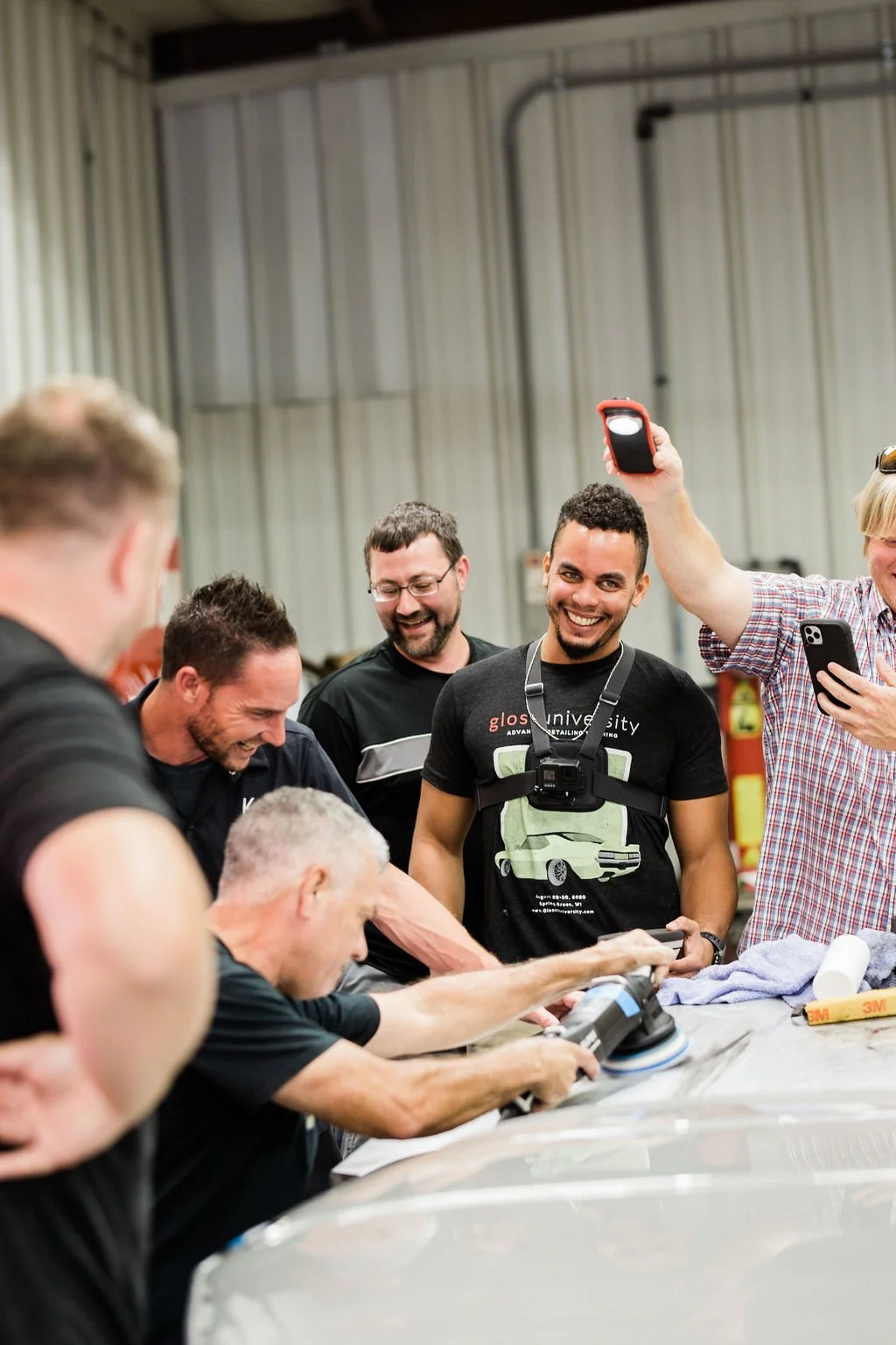 Group of men gathered around a car, smiling, with one man polishing the car's surface while others take photos and watch.
