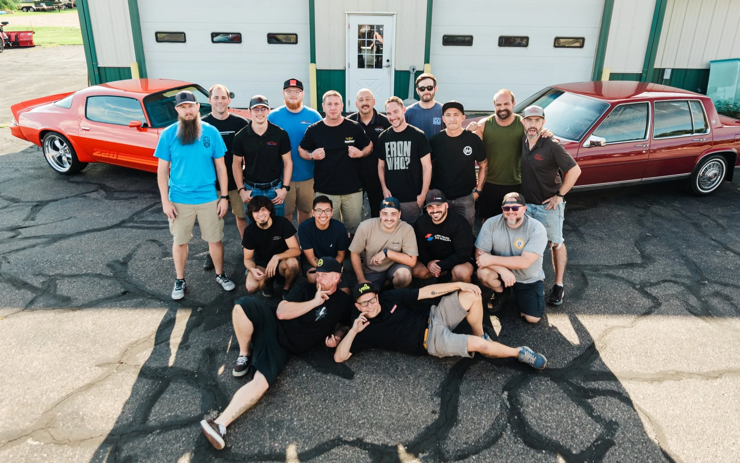 A group of 16 men gathered outdoors in front of a garage with two classic cars, one orange and one navy, on a cracked asphalt surface. The men are smiling and posing for the photo, with some sitting and others standing.