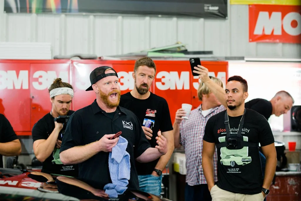 A group of people gathered indoors, with a man in the foreground speaking and gesturing, holding a phone and a towel, others listening with one taking a photo or video, in front of red 3M storage cabinets.