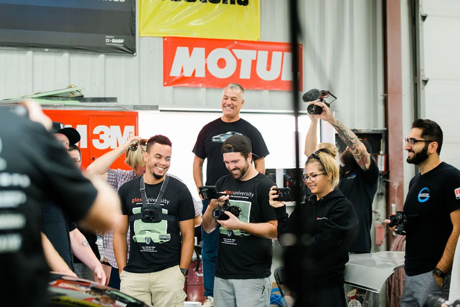 Group of people in a workshop, smiling and taking photos, with car-related posters on the wall.
