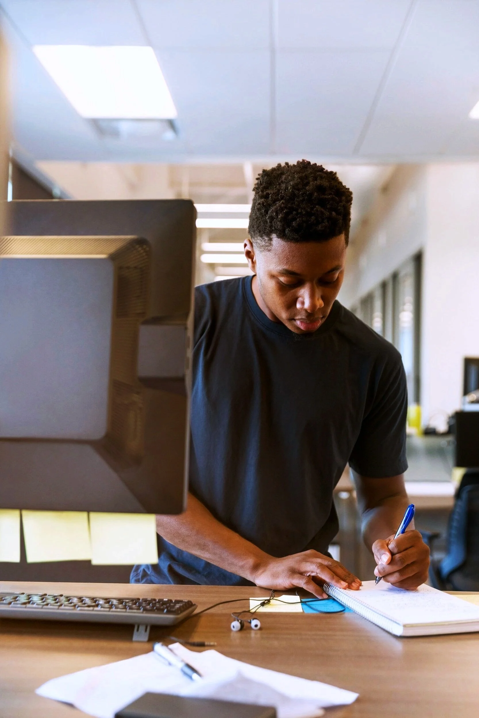 Young man standing at a desk writing in a notebook, computer monitor, keyboard, and earphones on the desk in an office setting.