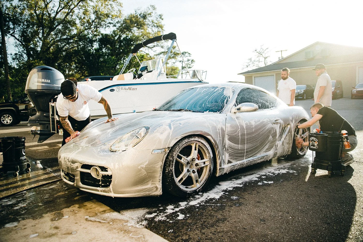 People washing a silver sports car with soap and water in a driveway, with a boat and parked cars in the background.