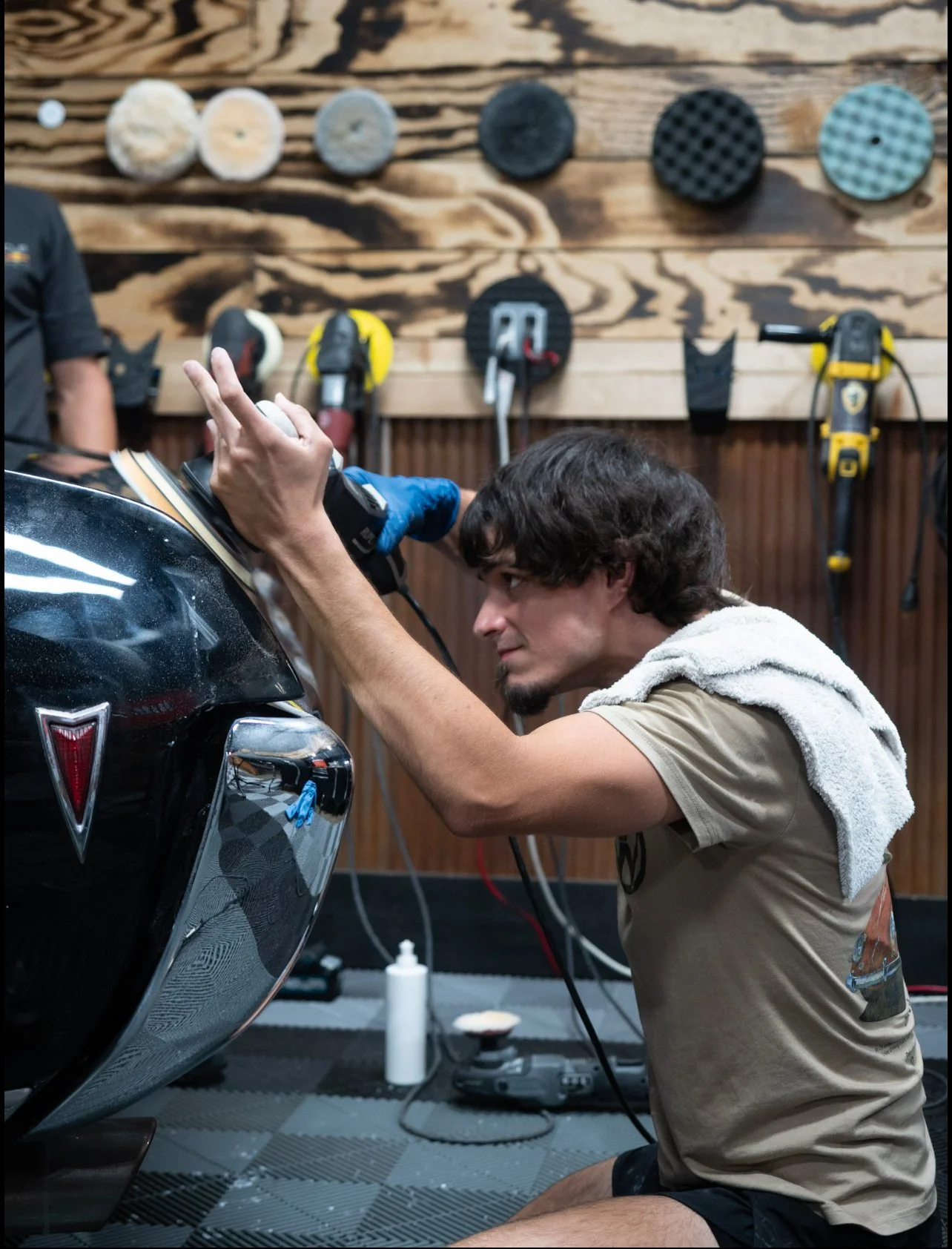 A man cleaning a classic black car at a garage, with wooden wall and car care tools hanging in the background.