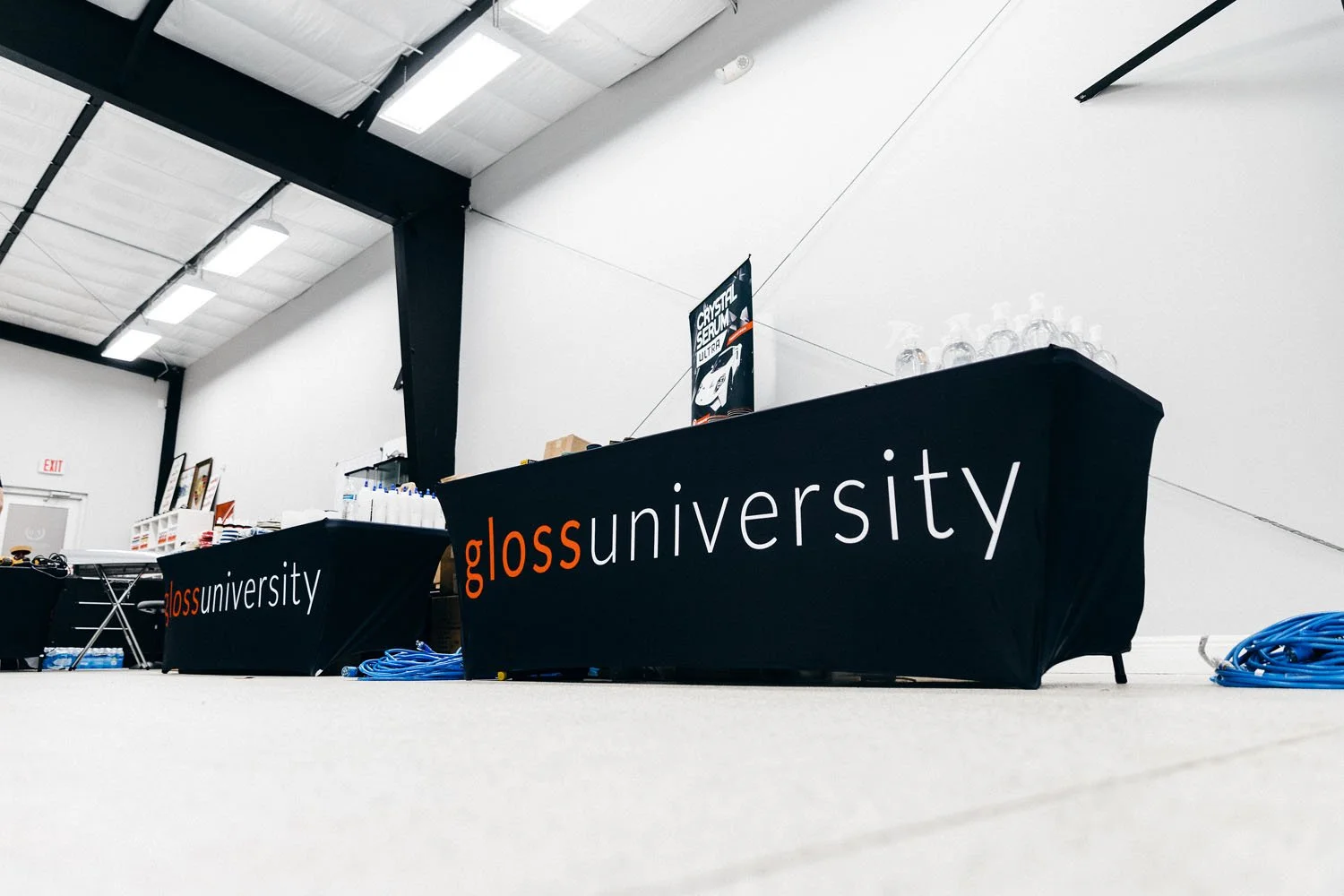 Tables covered with black cloths displaying 'gloss university' branding at an event space with white walls and ceiling, with water bottles and equipment on the tables.