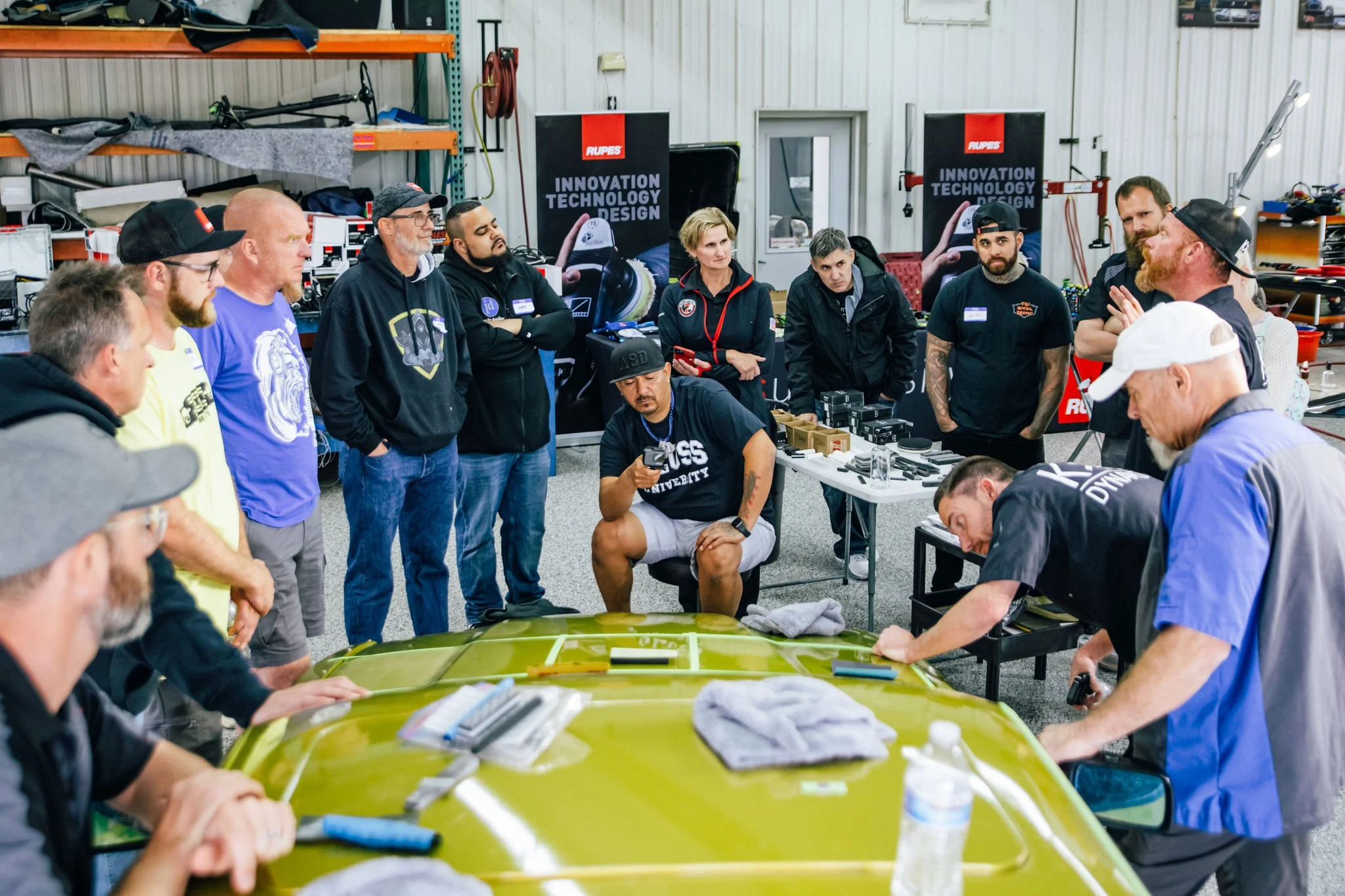 Group of people gathered around a car in a workshop, some working on the car, others observing, with banners in the background reading 'Innovation', 'Technology', 'Design', and various tools and equipment on tables.