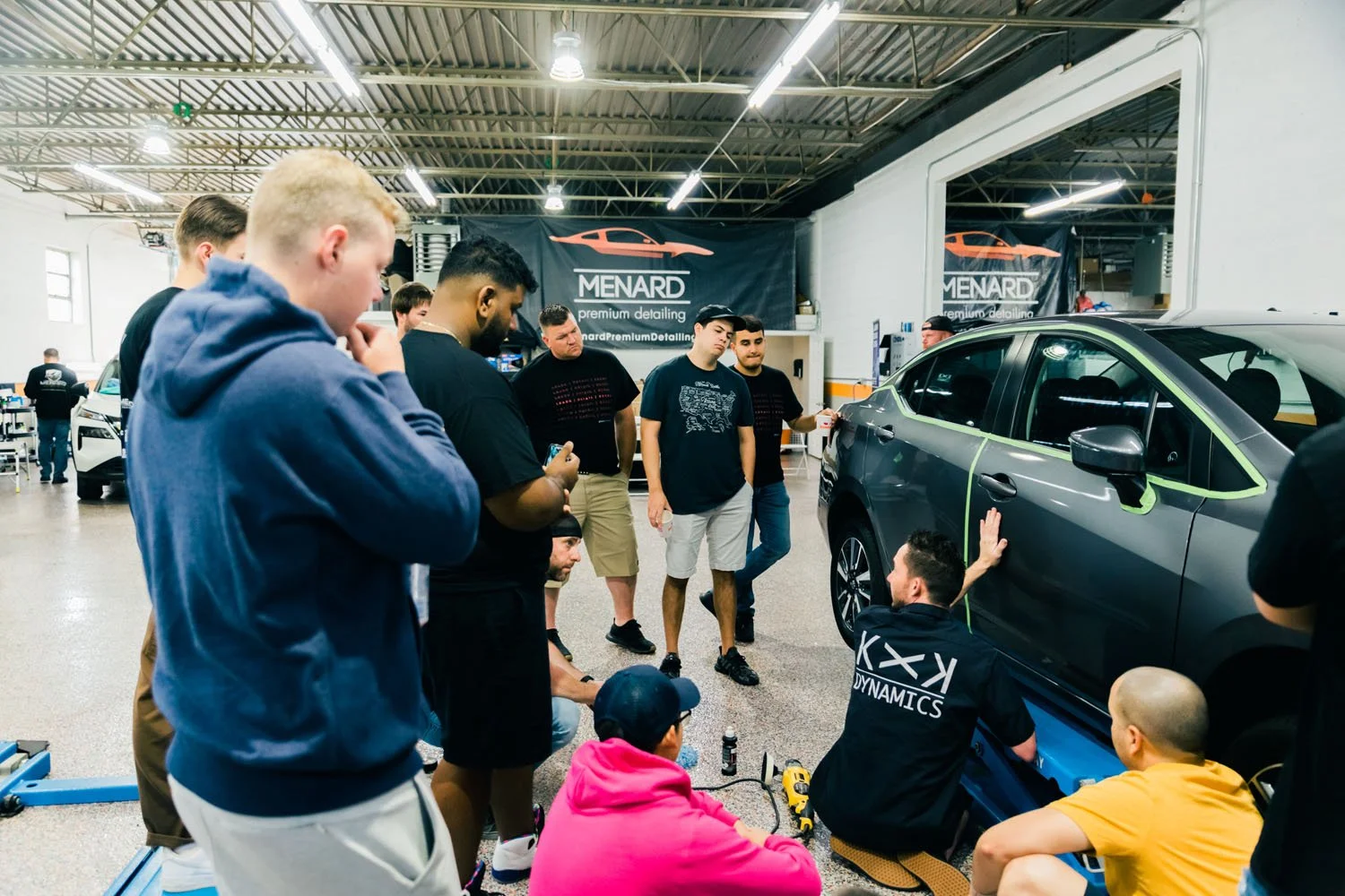 Group of people gathered around a car inside a detailing shop, where a man appears to be explaining or demonstrating something about the vehicle. The shop has bright indoor lighting and banners that read "MENARD premium detailing."