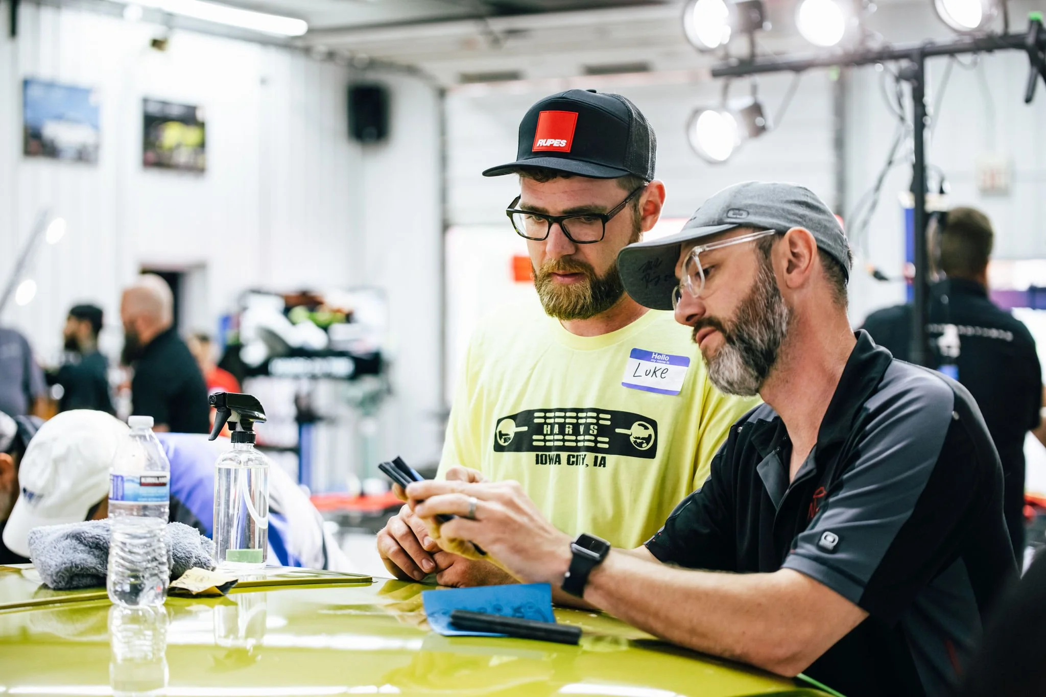 Two men with glasses and beards sitting at a yellow table, looking at a phone. The man on the left is wearing a yellow t-shirt with a graphic and a black cap with a red logo, and a name tag that says 'Luke.' The man on the right is wearing a gray cap
