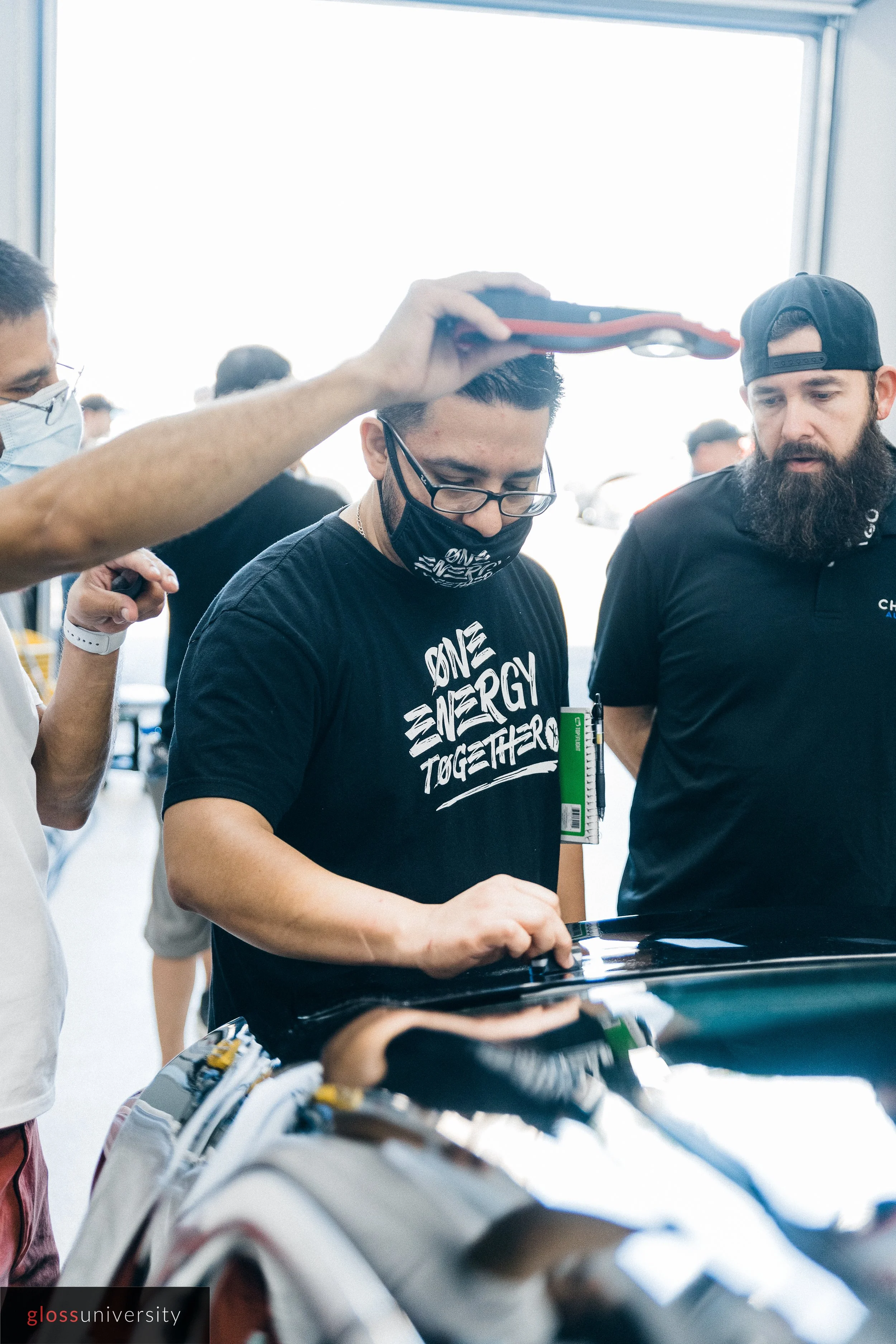 A group of people gathered around a car, with one person in the center inspecting the vehicle while another person holds a scanner above him. All individuals are wearing masks.